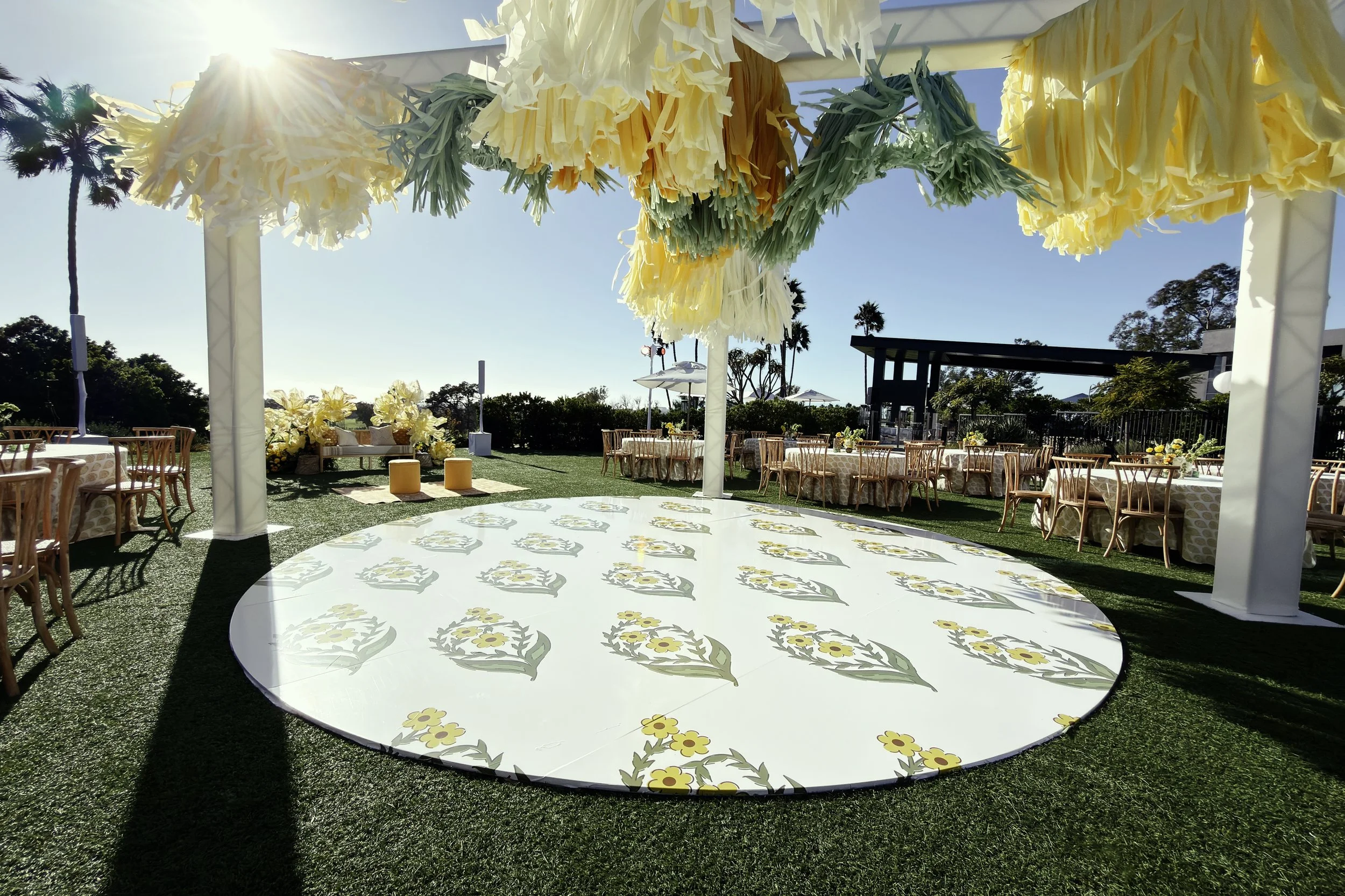 Outdoor event setup with decorated tables and floral decorations, under a canopy with hanging floral arrangements, in a sunny area with palm trees and a blue sky.