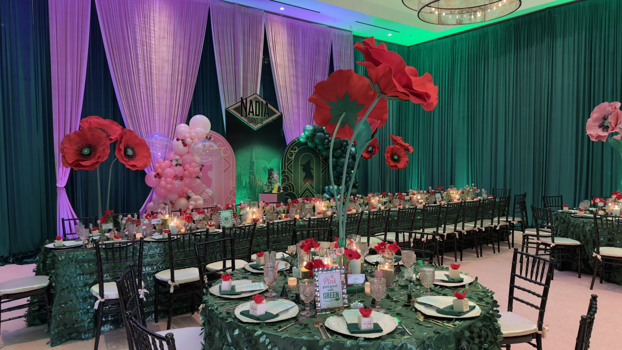 Decorated banquet hall for a celebration with a backdrop of purple, pink, and green curtains, large red and pink floral centerpieces, balloon arrangements, and a long table set with green tablecloths, candles, and flower accents.