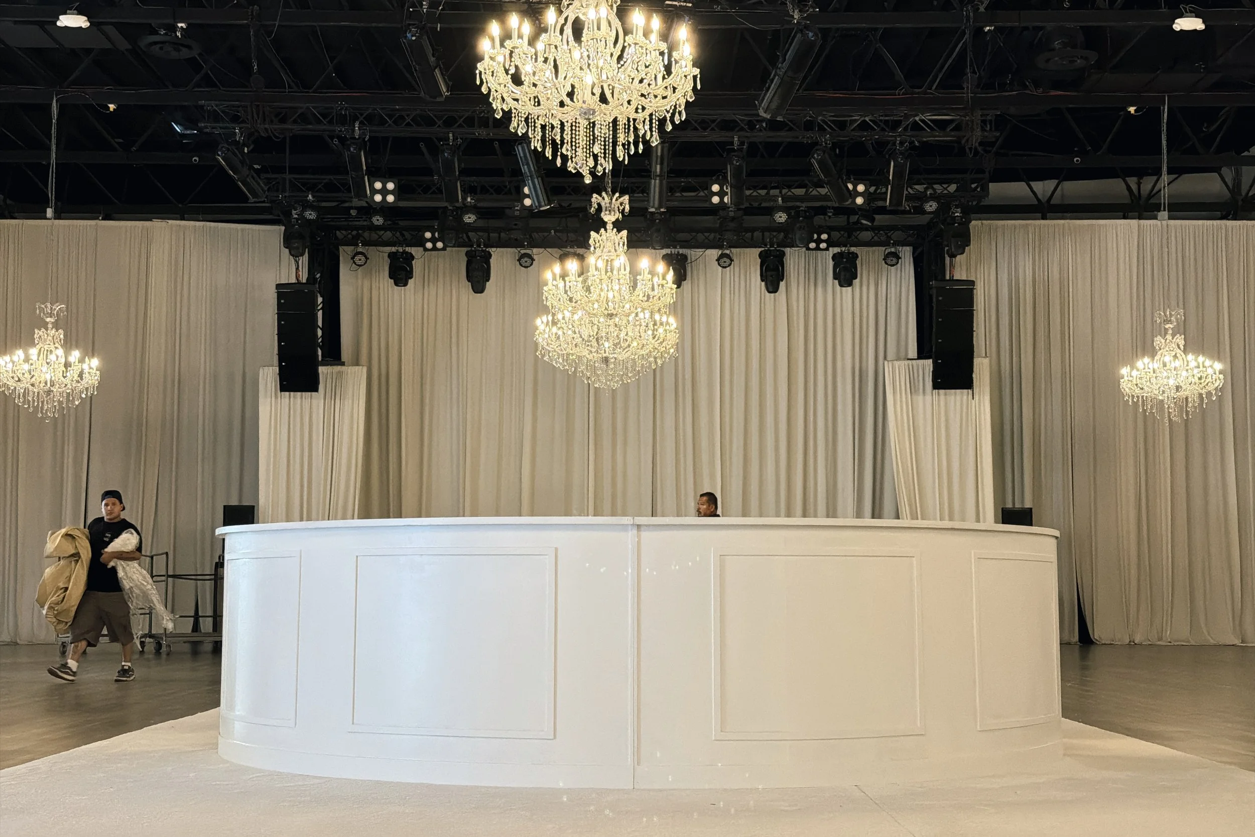 Empty stage with a white booth or bar in front, decorated with multiple chandeliers hanging from the ceiling. The backdrop is draped with white curtains. Two men are present to the left of the stage, one holding a large object and the other standing 