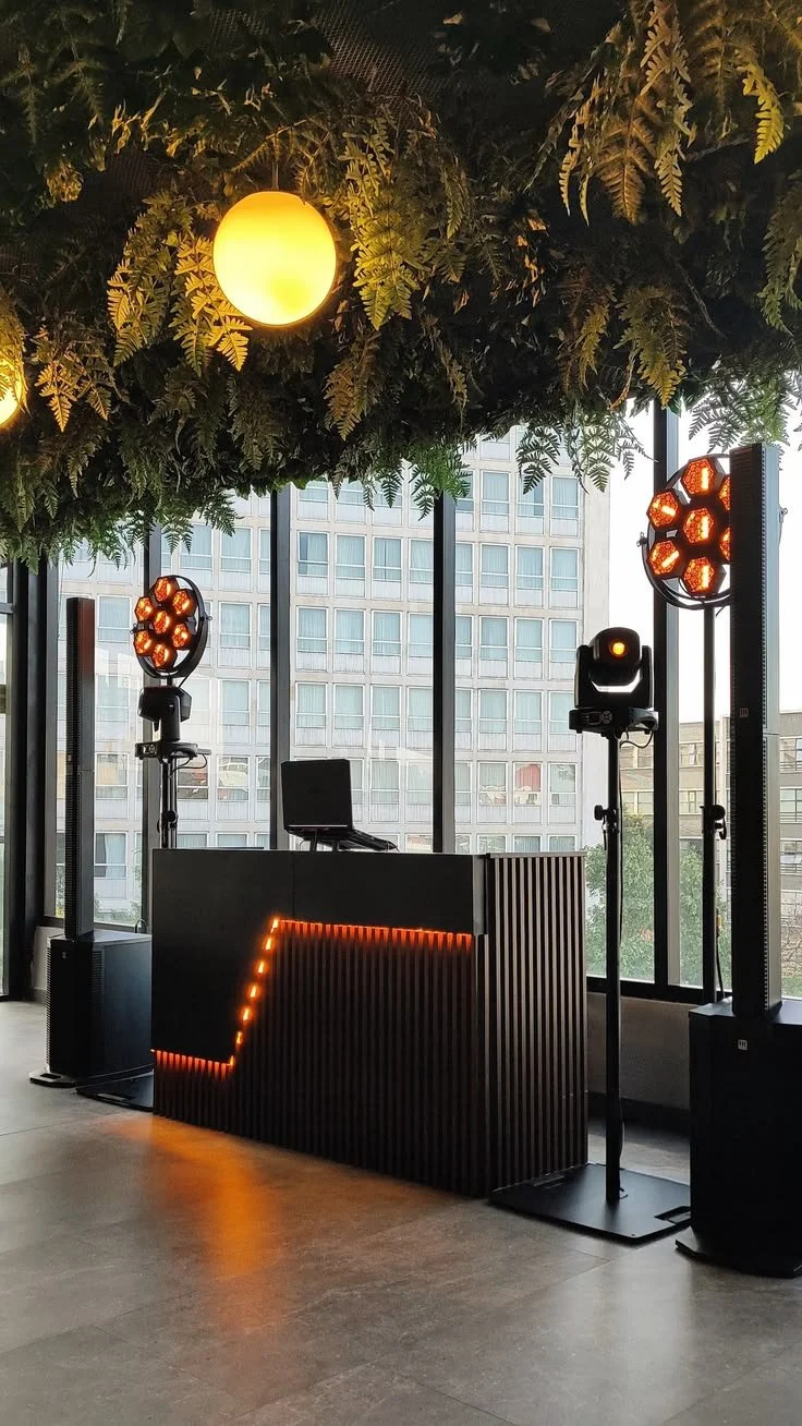 Indoor event space with DJ setup, including two speaker lights, a DJ booth with LED lighting, and a leafy ceiling decoration with warm yellow lights, large glass windows showing a cityscape.