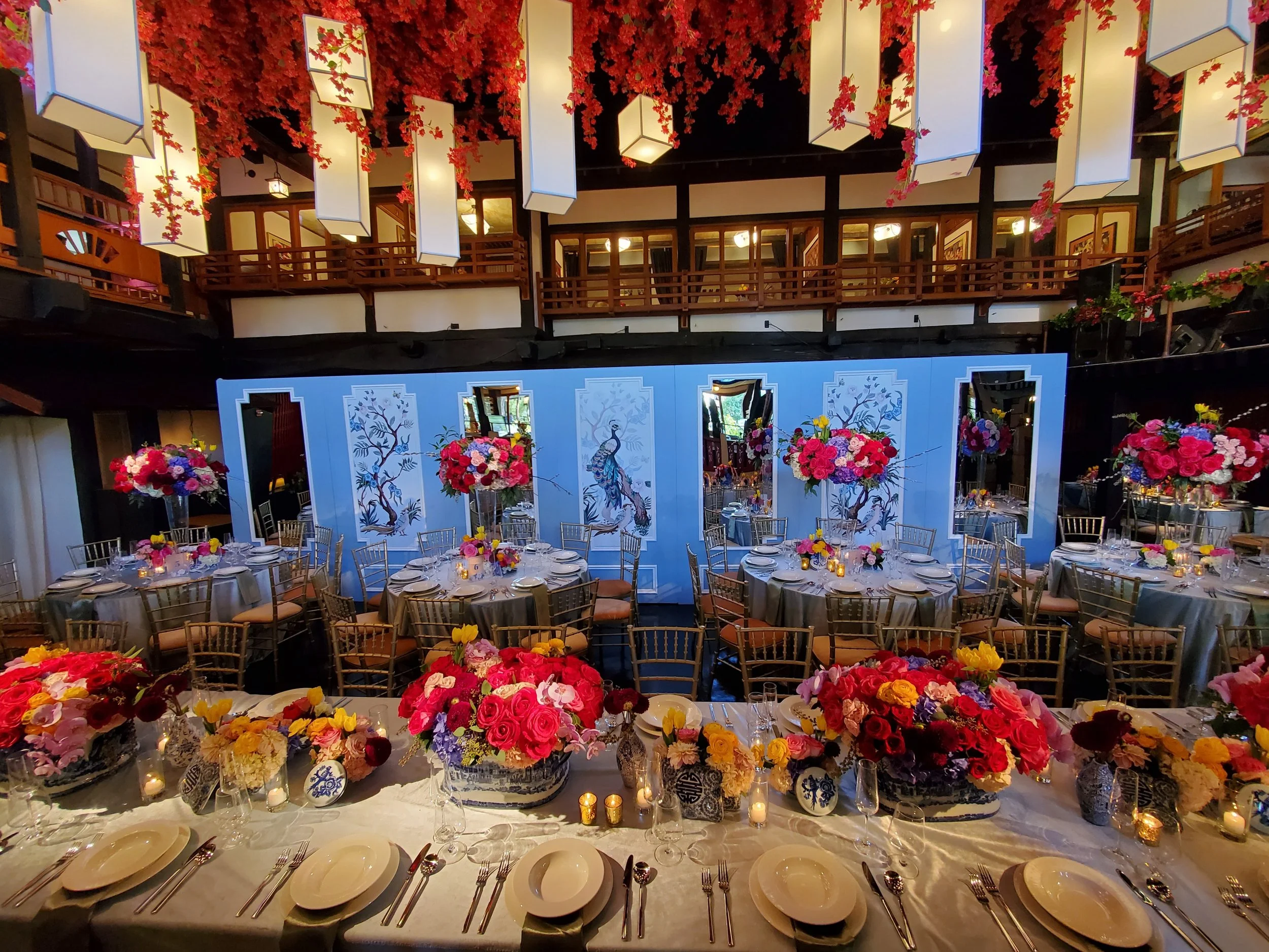 Elegant event space set for a banquet with round tables and floral centerpieces, a decorated wall with peacock art, and a balcony with hanging decor and red floral arrangements.