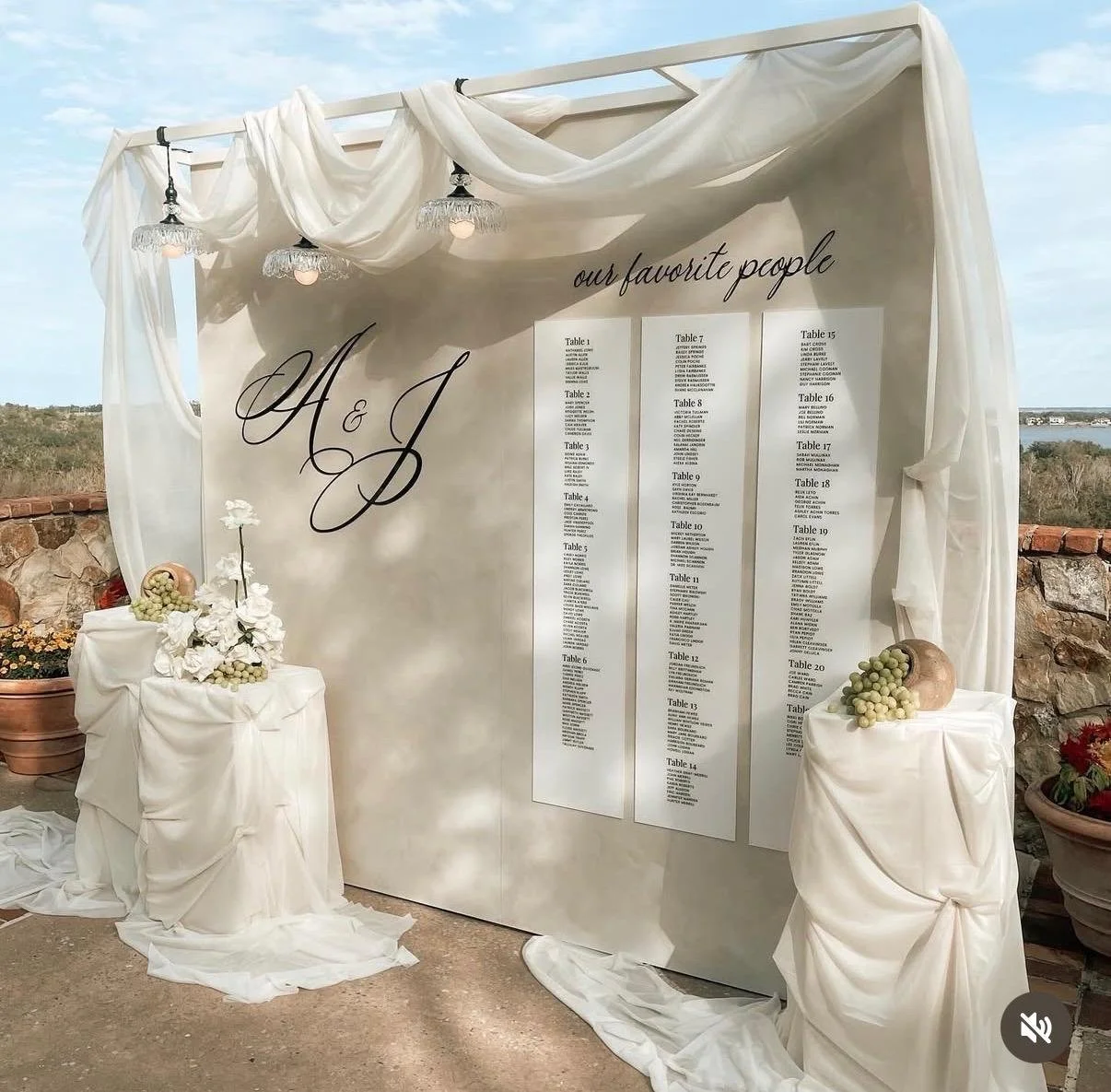 Wedding seating chart display with white drapery, chandeliers, and floral decorations, listing tables and guest names.