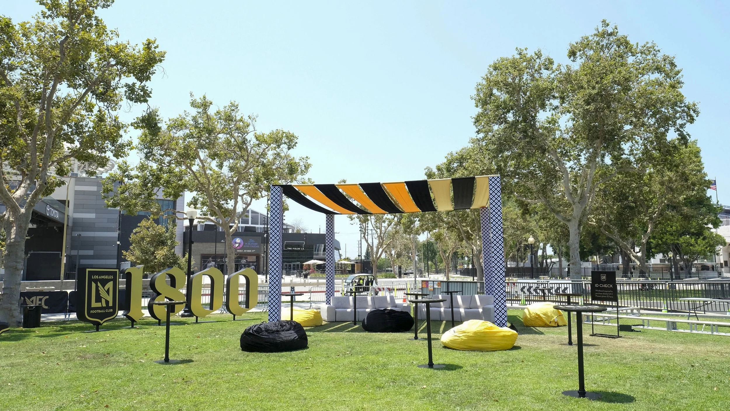 An outdoor seating area in a park with trees, yellow bean bags, chairs, and decorative arches, with a stadium or arena in the background.