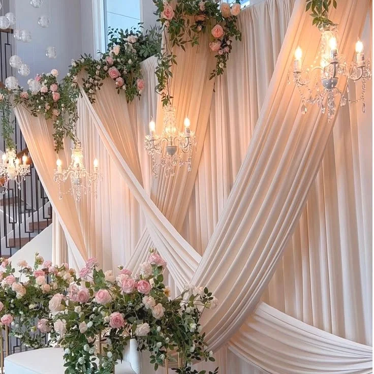 Elegant wedding backdrop featuring flowing white and blush curtains, pink and white rose floral arrangements, and crystal chandeliers.