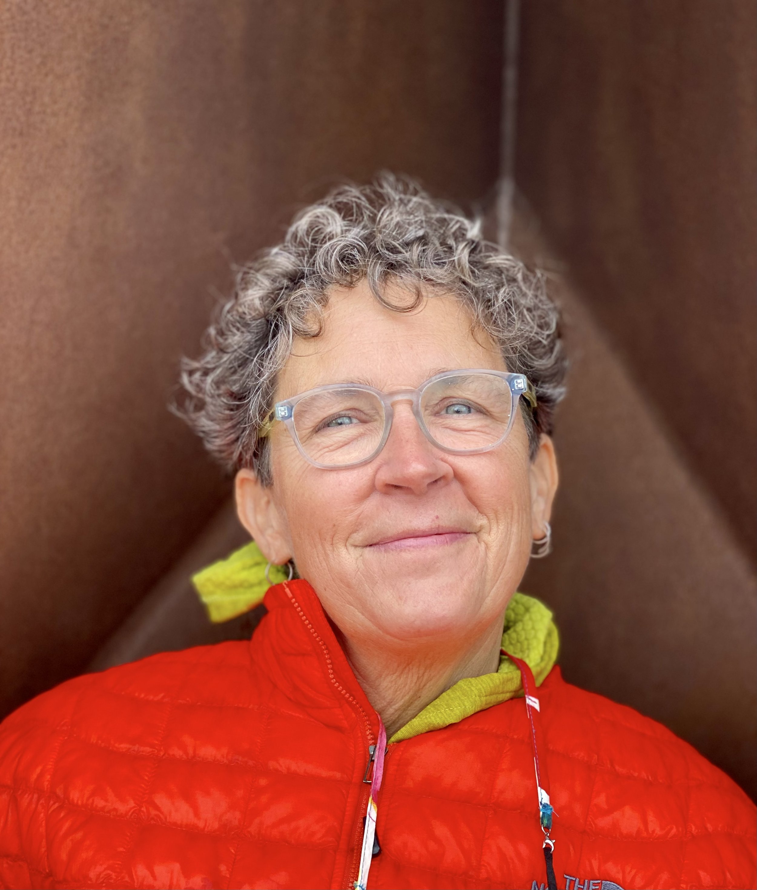 Close-up of a smiling woman with curly gray hair, wearing clear eyeglasses, a red jacket, and a yellow hoodie, against a brown background.