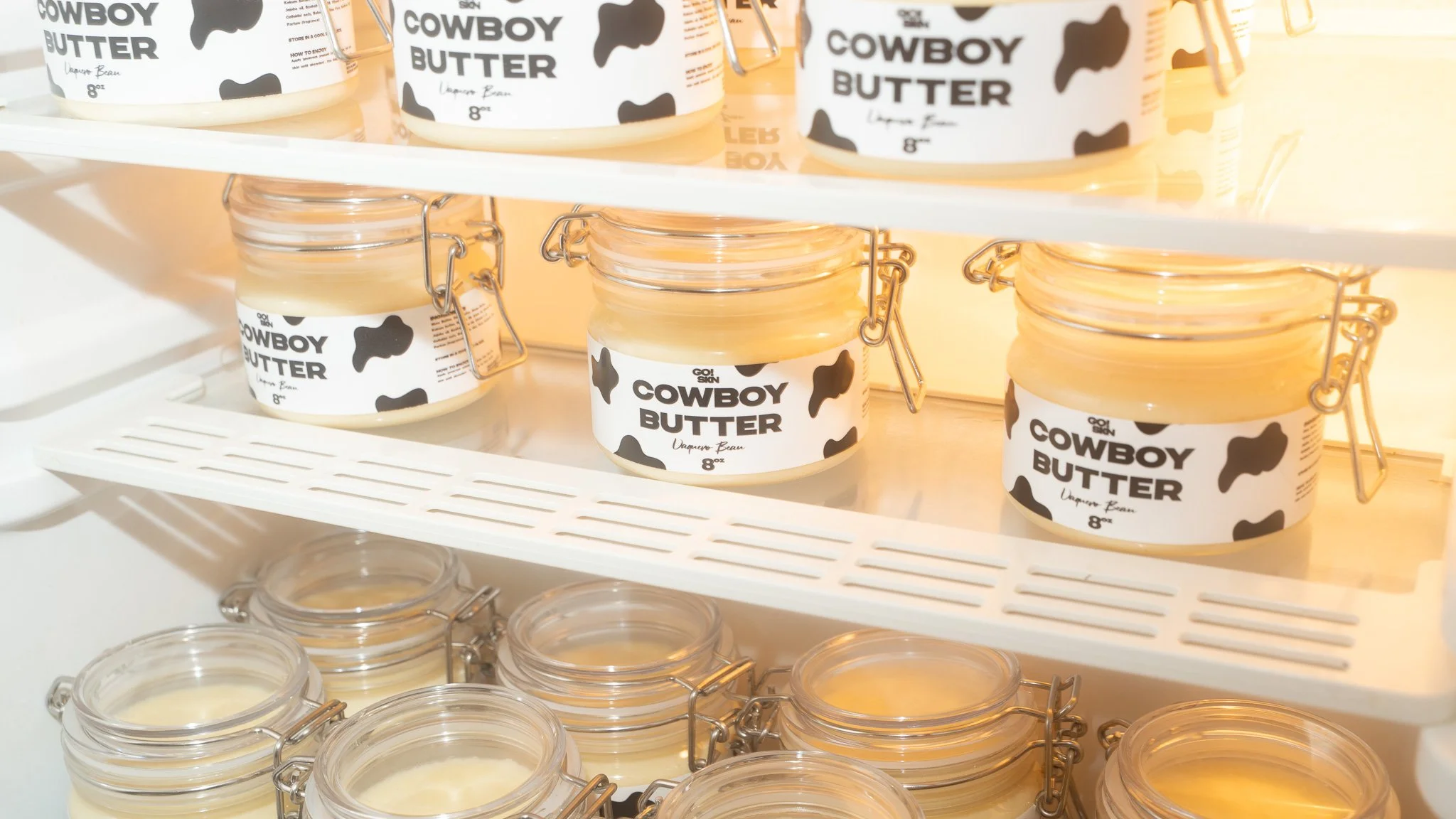 Shelf displays multiple jars of Cowboy Butter with black and white cow print labels, some open with lids placed below, in a retail setting.