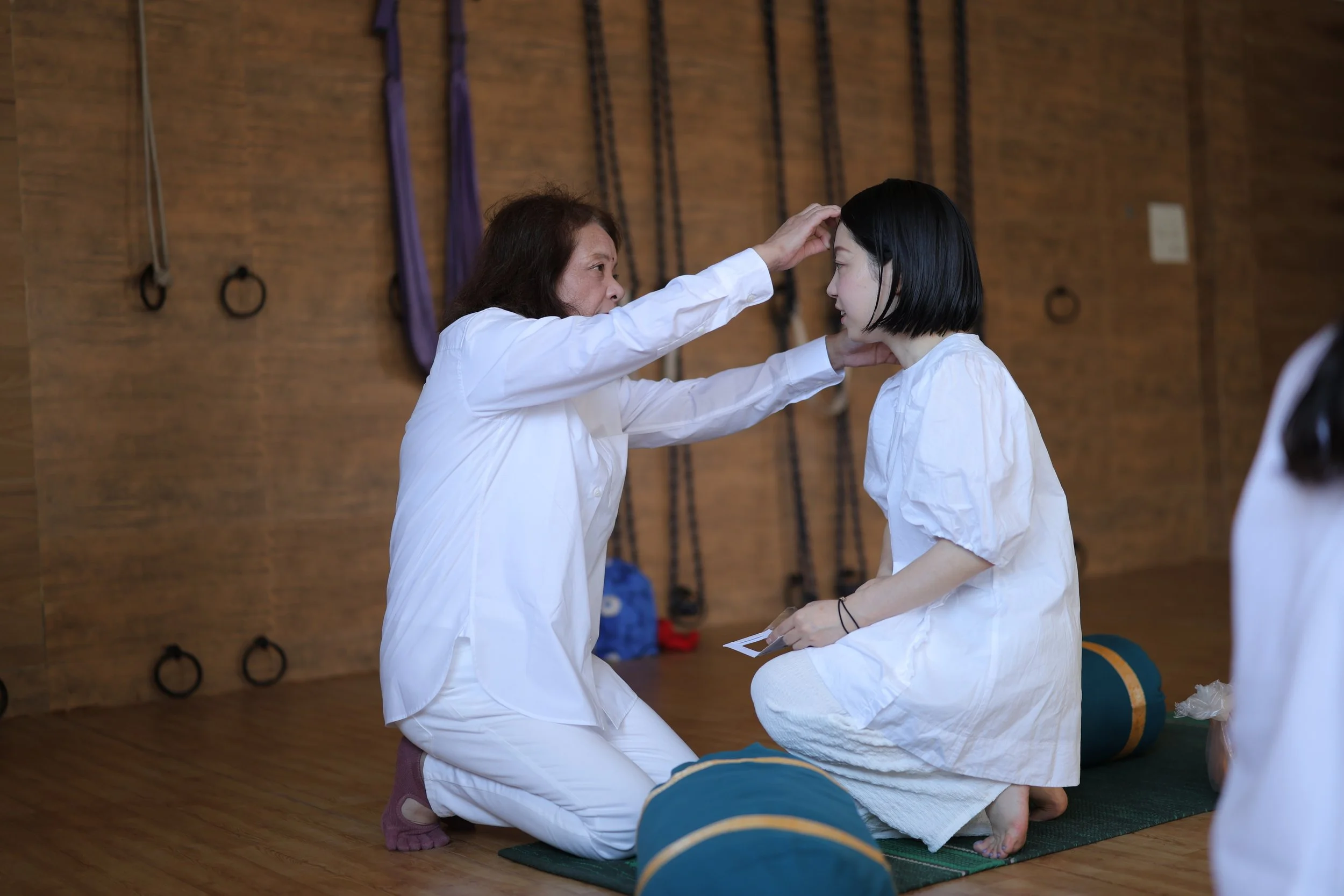 Three people practicing yoga on a platform outdoors with trees, a blue sky, and buildings in the background. Two women and one man in meditation pose with hands together in prayer position.