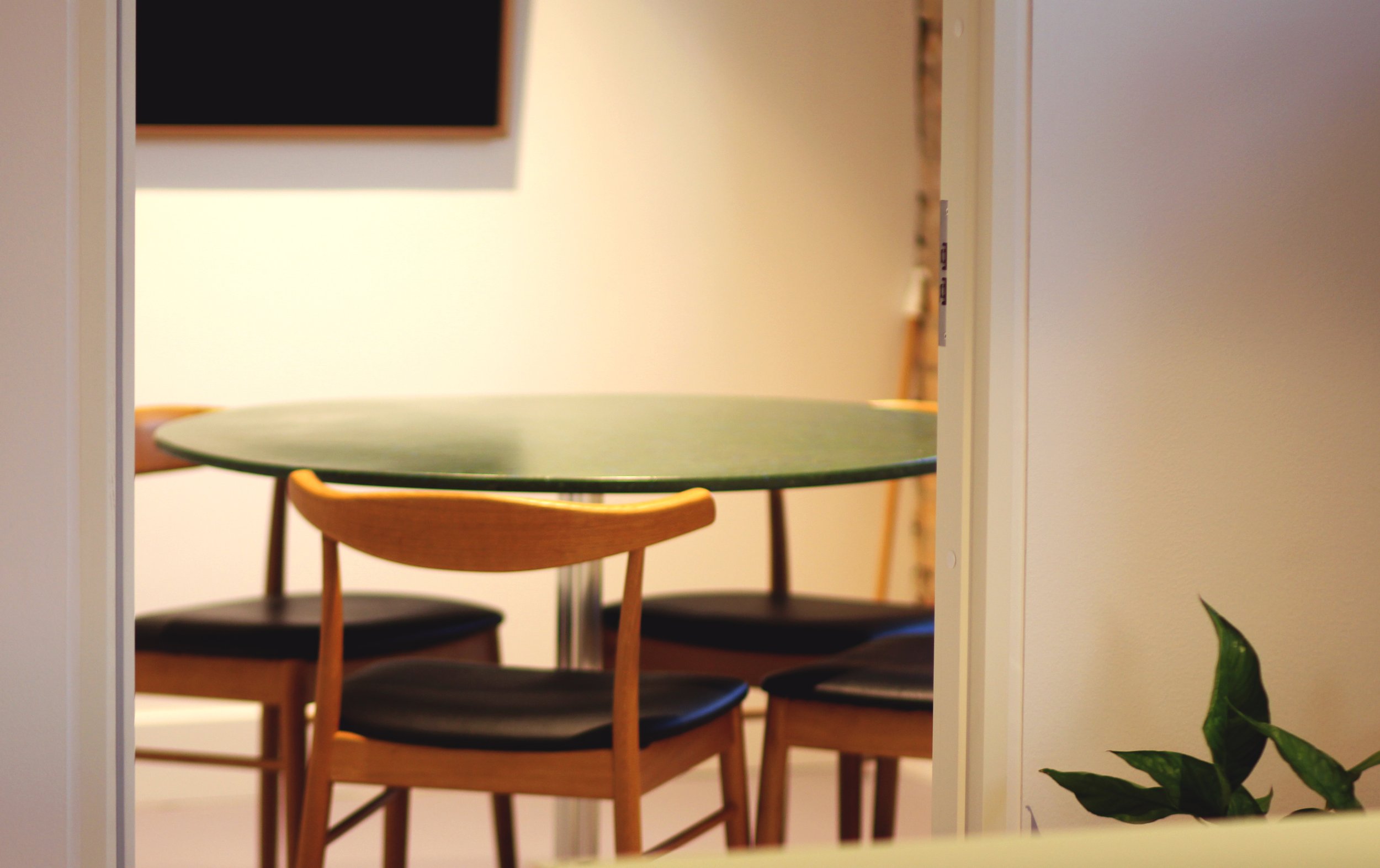 A round green table with four wooden chairs, viewed through a doorway in a room with a plant in the foreground.