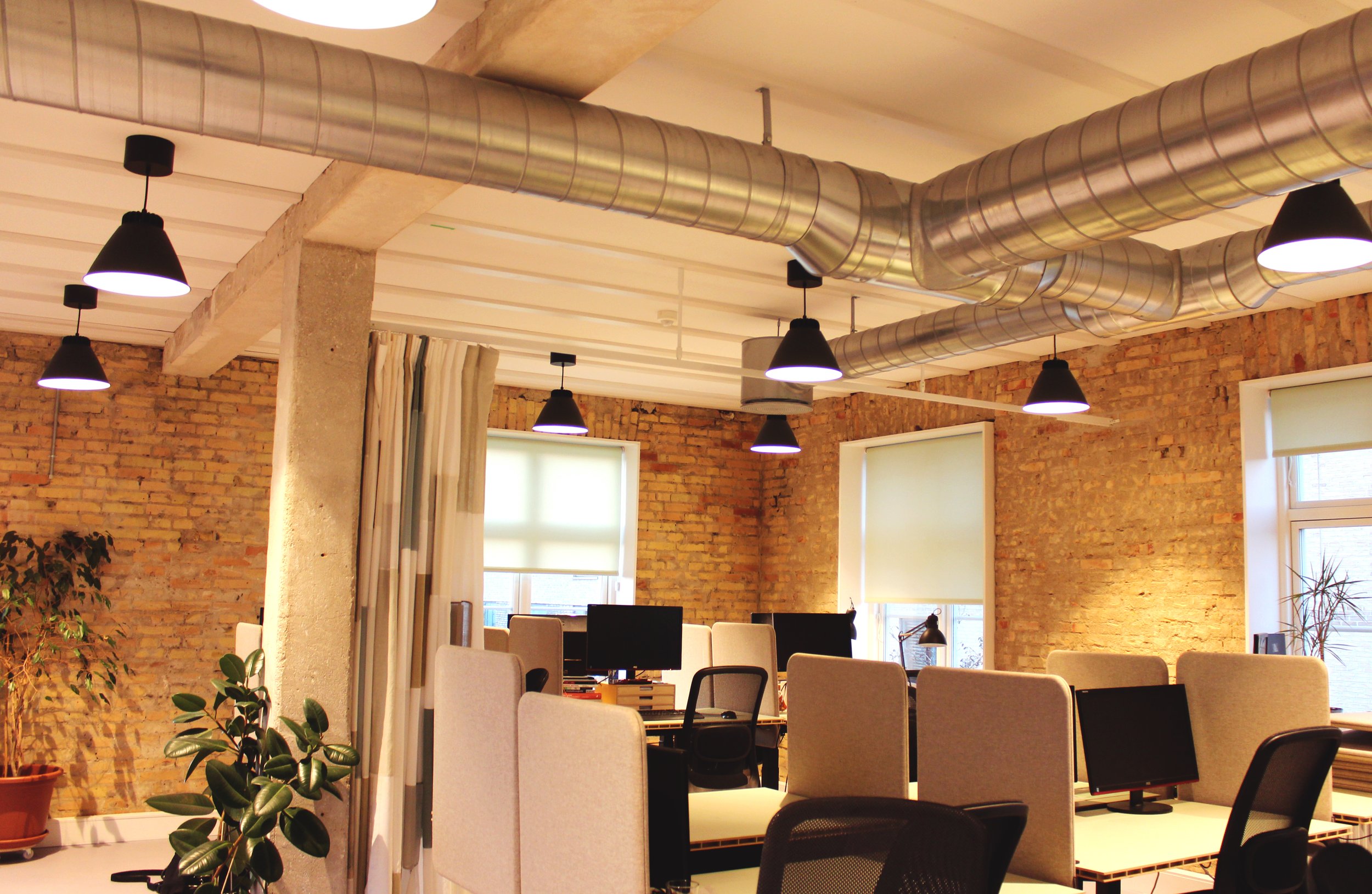 An office space with exposed brick walls, several desks with computers, and black pendant lights hanging from the ceiling. There are large windows with blinds, potted plants, and a curtain partially covering one window.