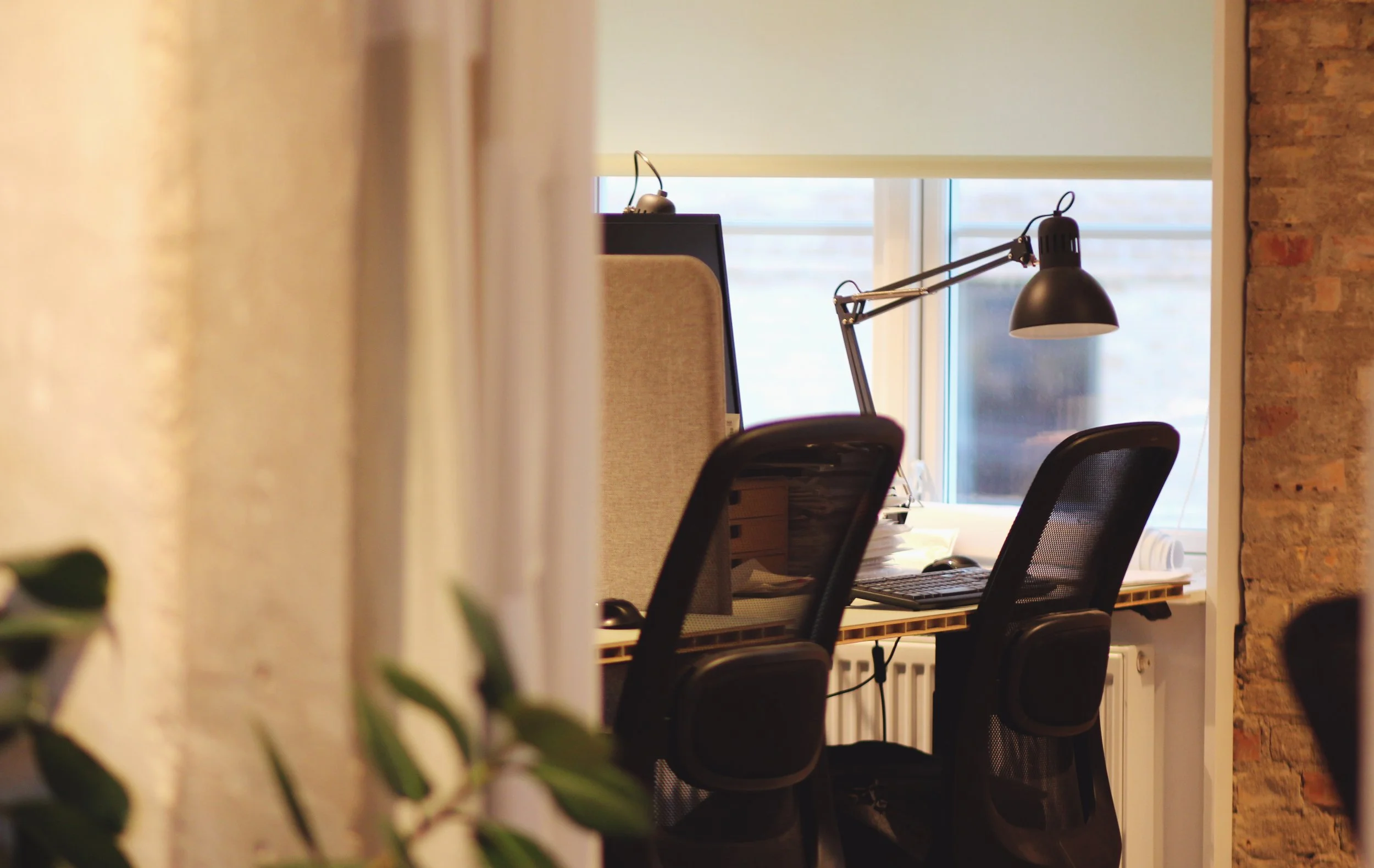 An office workspace with chairs, a desk, a computer monitor, and a desk lamp by a window, with a brick wall on one side.