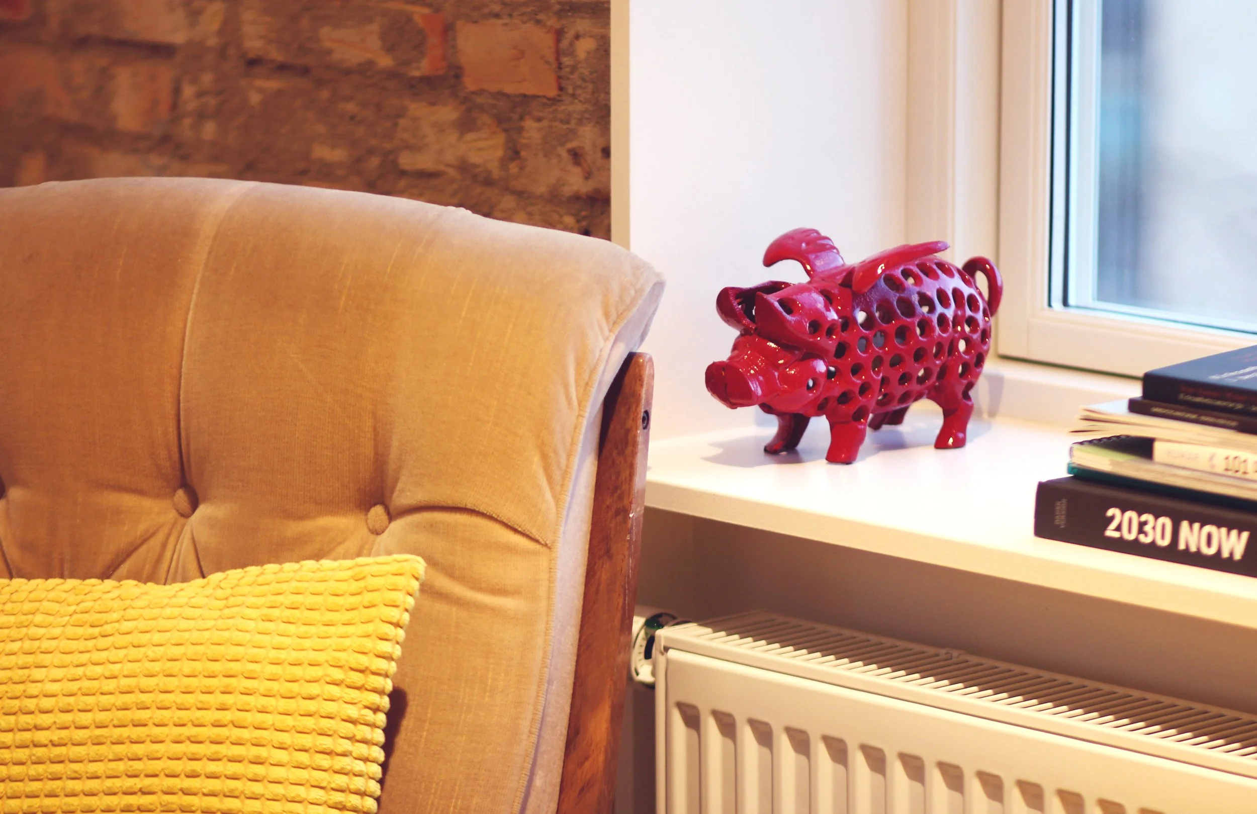 Decorative red pig-shaped sculpture with holes on a white window sill, next to stacked books and a beige armchair with a yellow textured pillow.