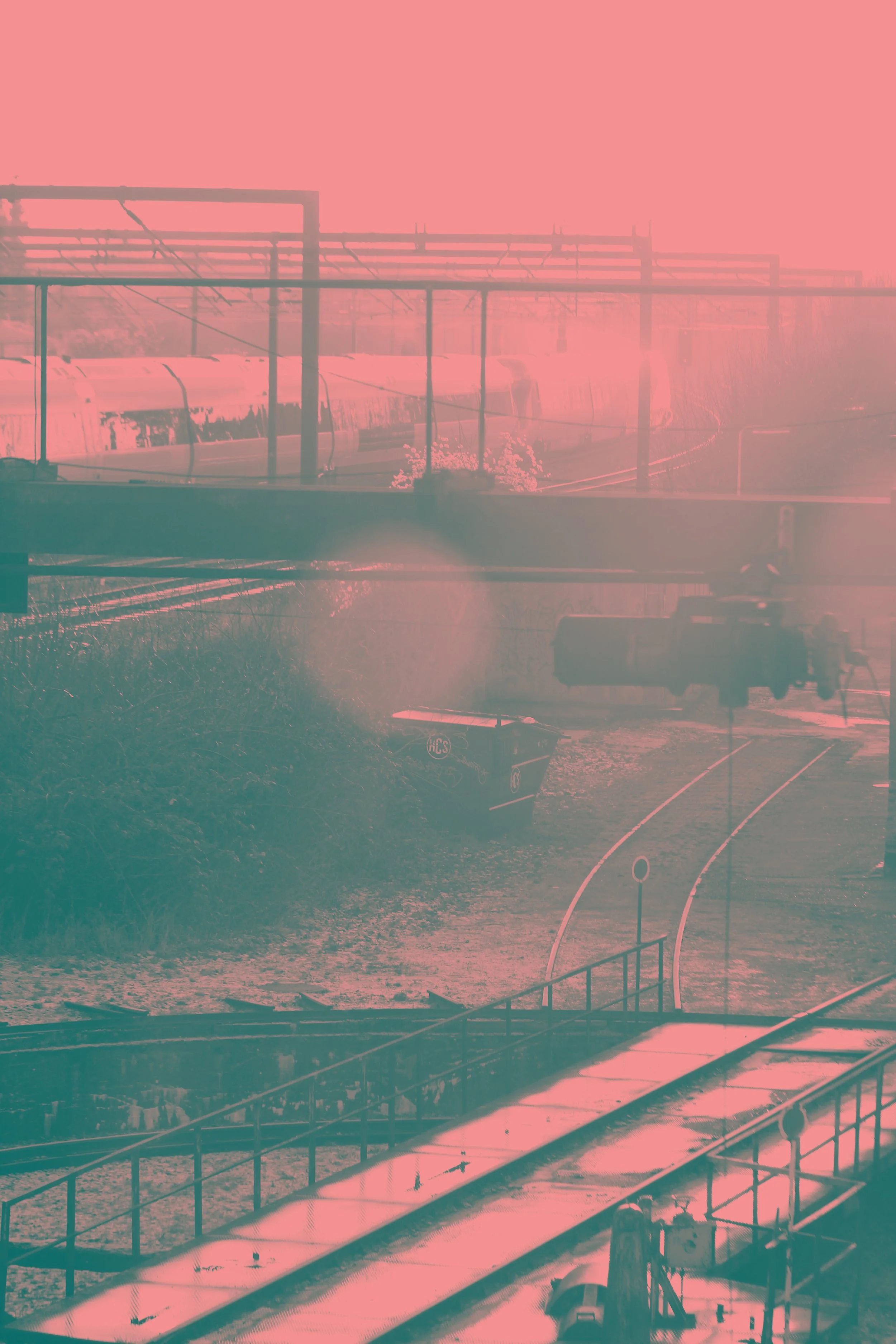 Train tracks with a passing train and a trash bin near the tracks, overpass and utility poles in the background, sunlight creating a pinkish hue.
