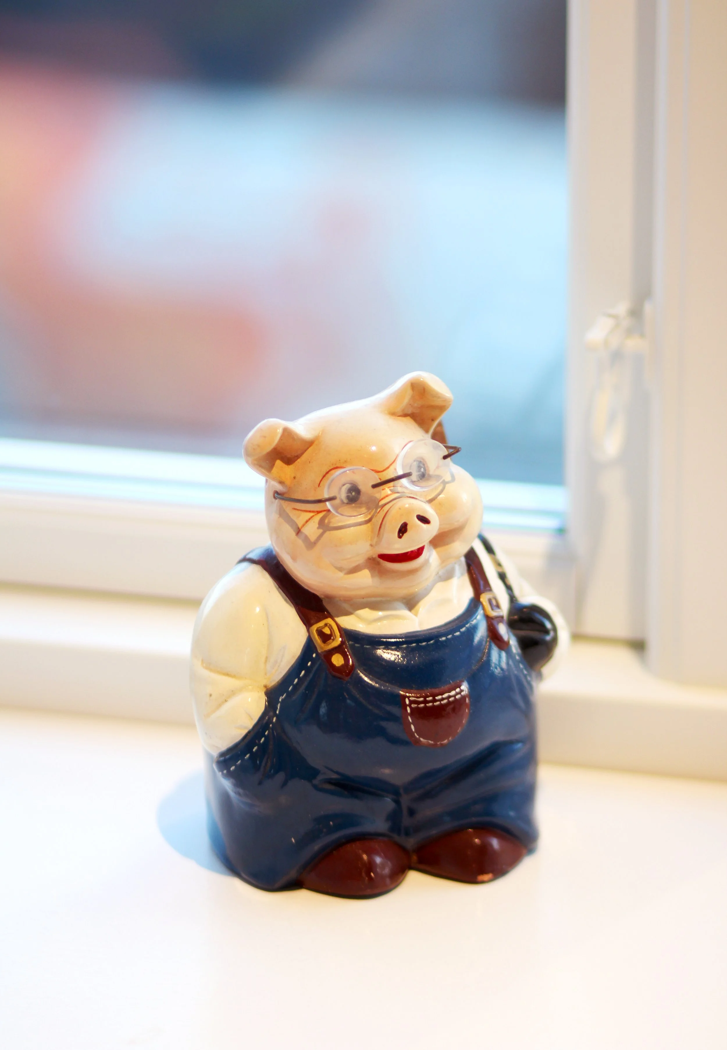 A pig-shaped ceramic figurine wearing glasses, a yellow shirt, blue overalls, and brown shoes, standing on a white windowsill near a window.