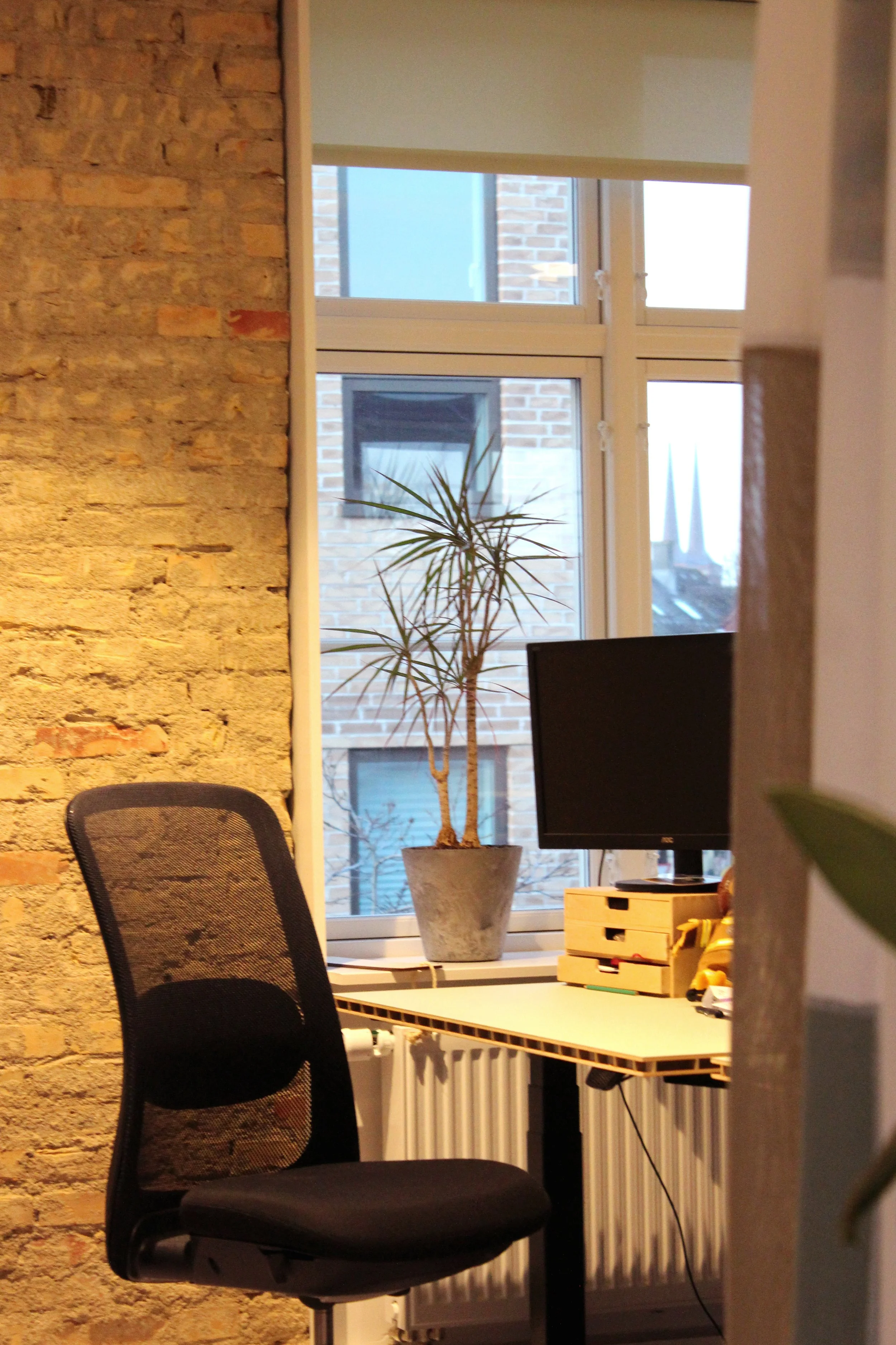 An office space with a brick wall, a black ergonomic chair, a desk with a computer monitor, a potted plant, and a window showing a brick building outside.
