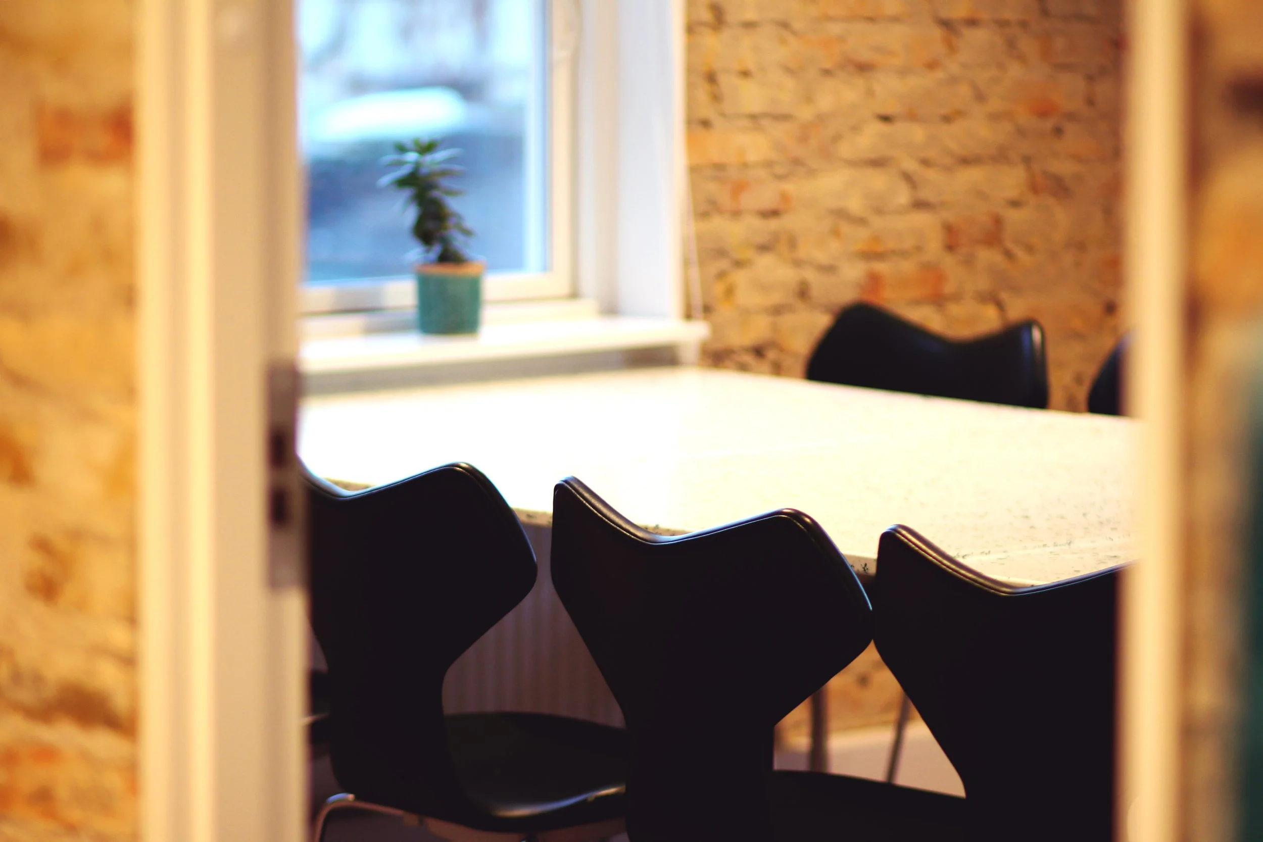 A view of a conference room through a partially open door. The room has a white table with black chairs around it, a window with white curtains, and a small potted plant on the windowsill. The walls are brick.