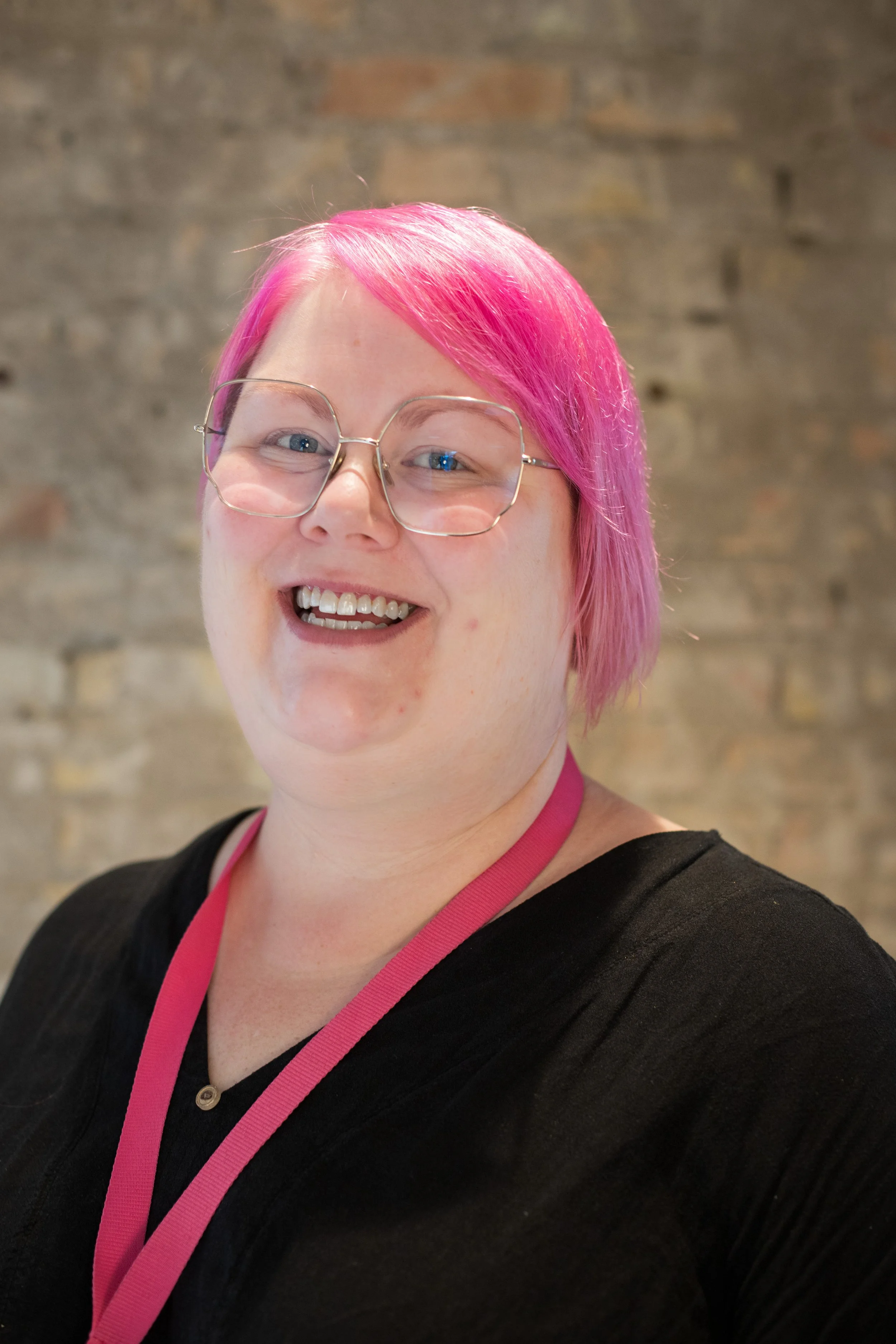 A woman with pink hair, glasses, and a black top smiling at the camera, wearing a pink lanyard against a blurred brick wall background.