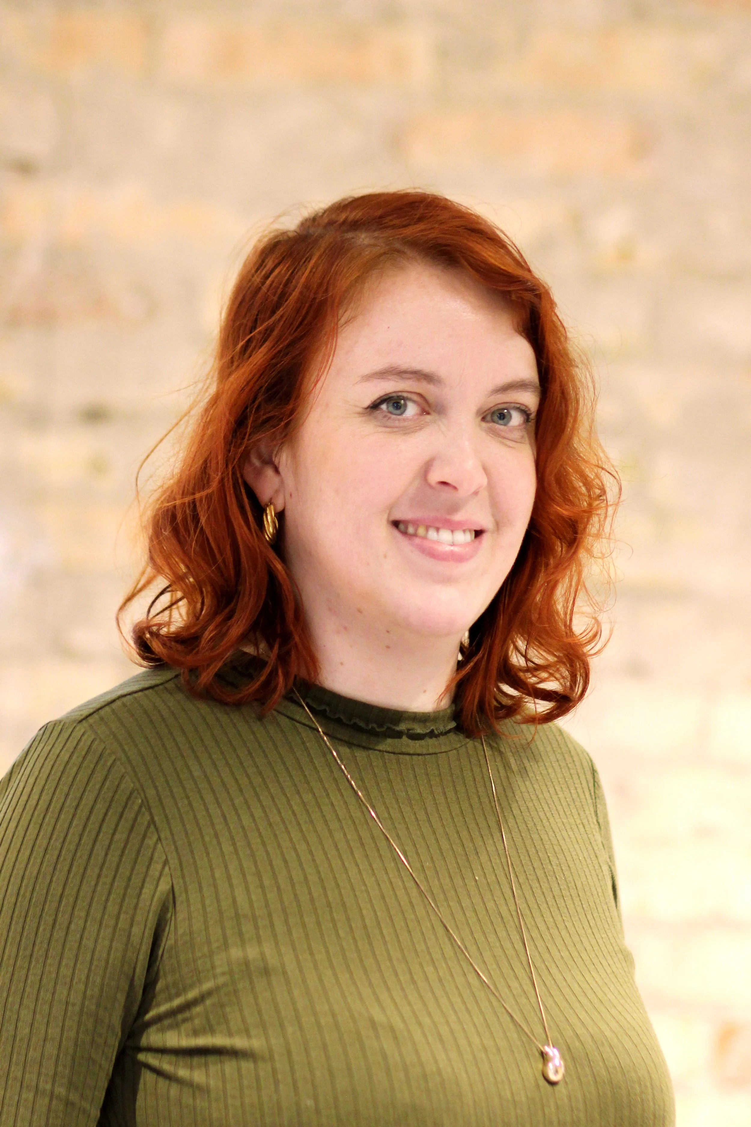 A woman with shoulder-length red hair, wearing a green top and gold jewelry, smiling in front of a brick wall.