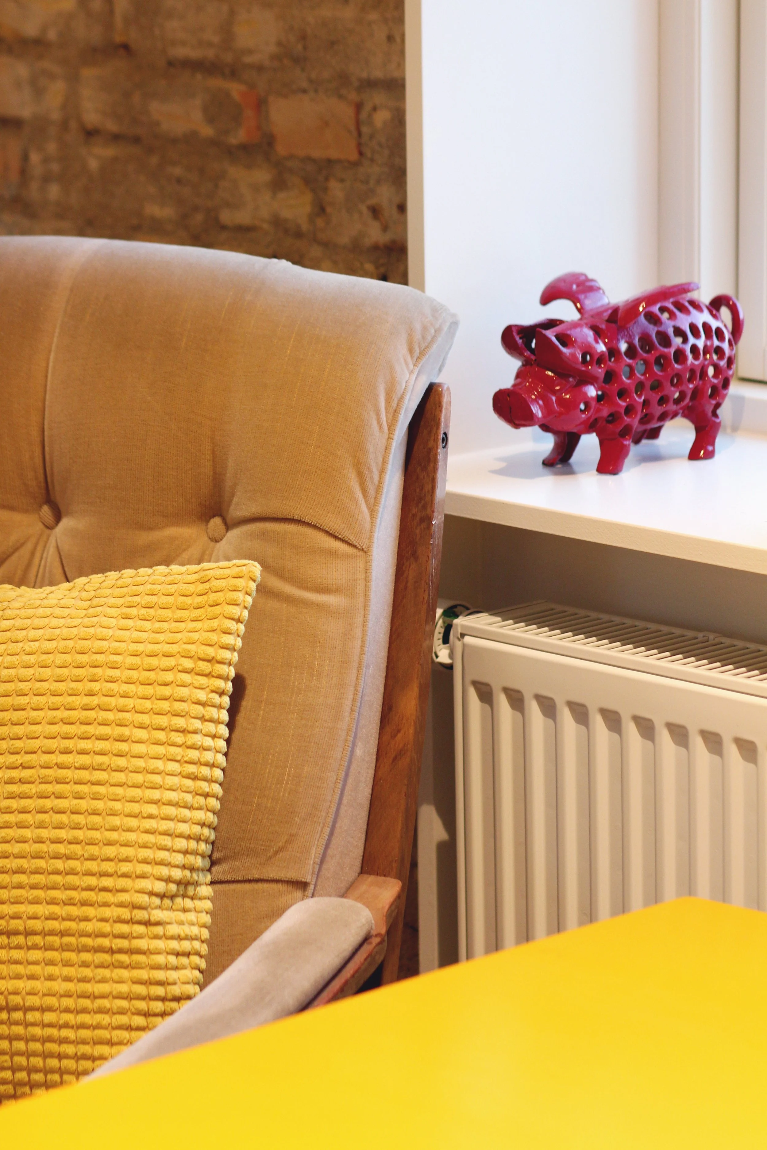 Corner of a room with a beige upholstered couch, a yellow textured pillow, a red ceramic pig-shaped sculpture with holes, a white radiator, and a windowsill.