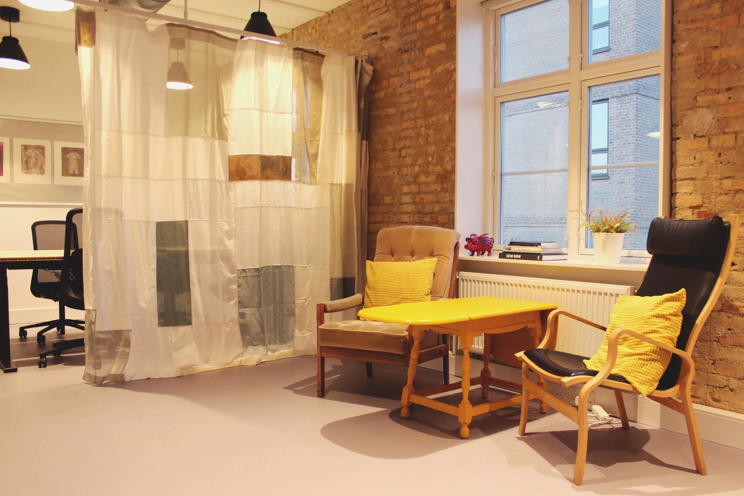 Cozy corner of an office with two armchairs, a yellow side table, and a window with brick exterior visible, separated by a fabric curtain.