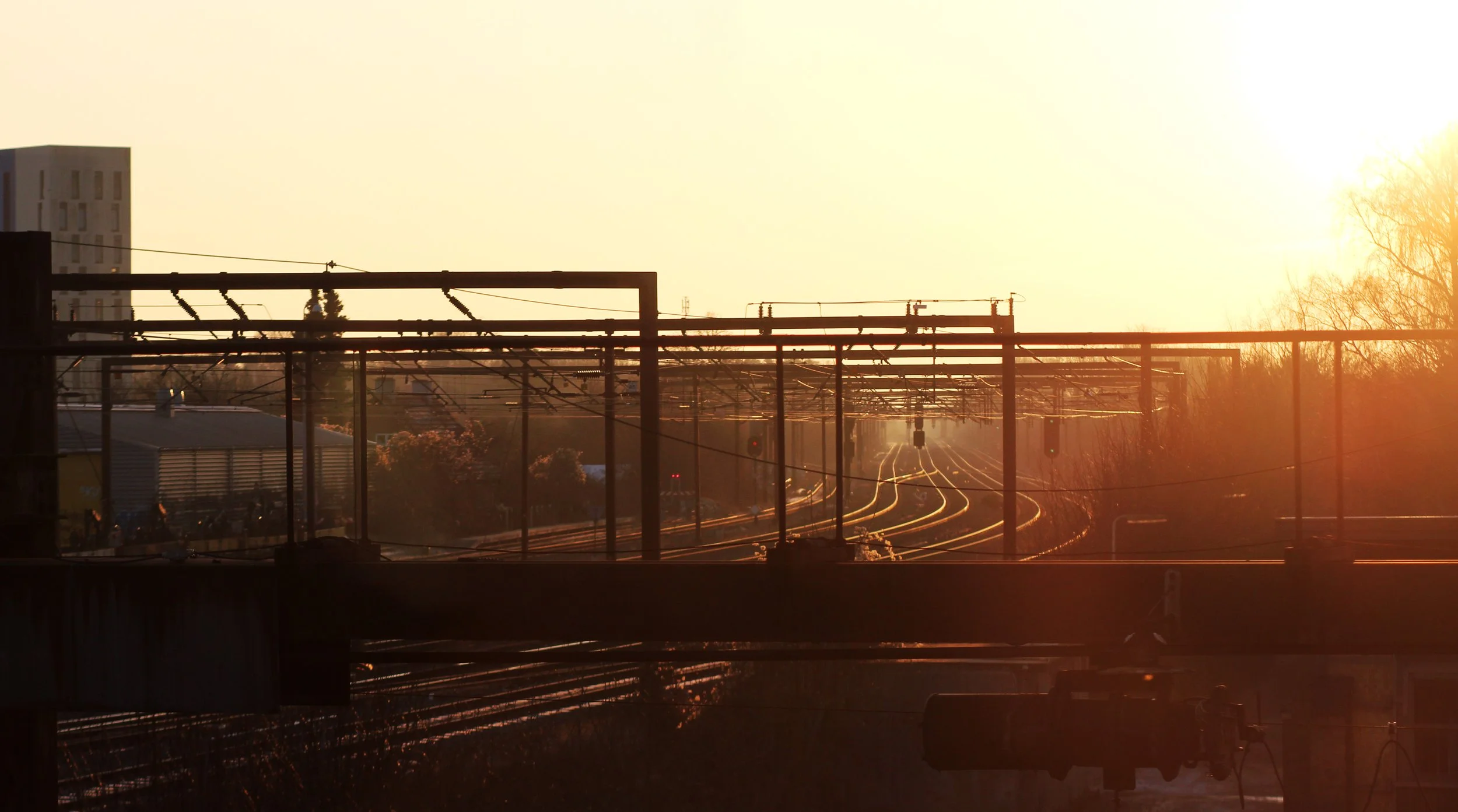 Train tracks extending into the distance with overhead electrical wires, illuminated by warm sunlight at sunset.