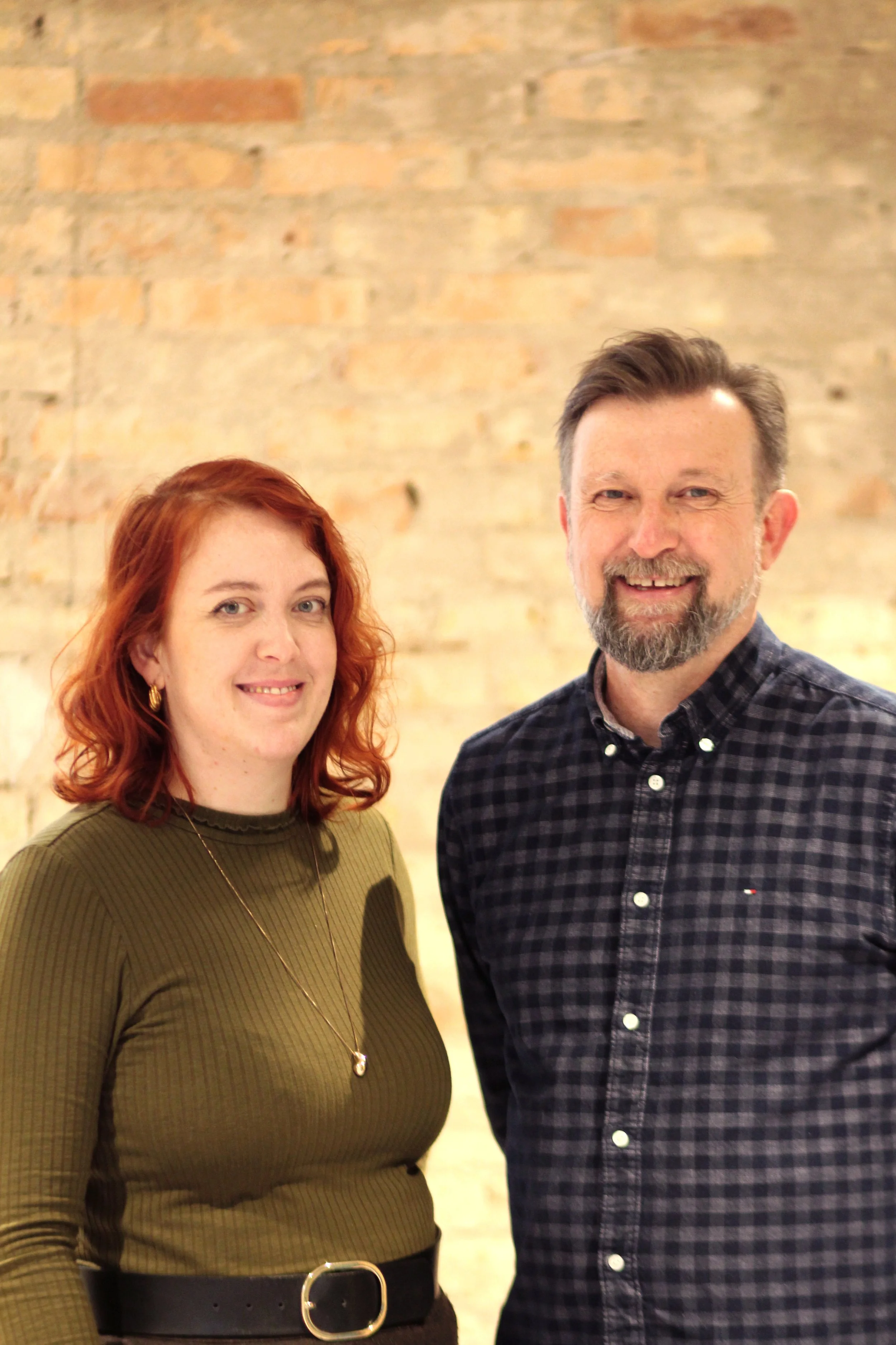 A woman with red hair and a man with a beard standing in front of a brick wall, smiling at the camera.