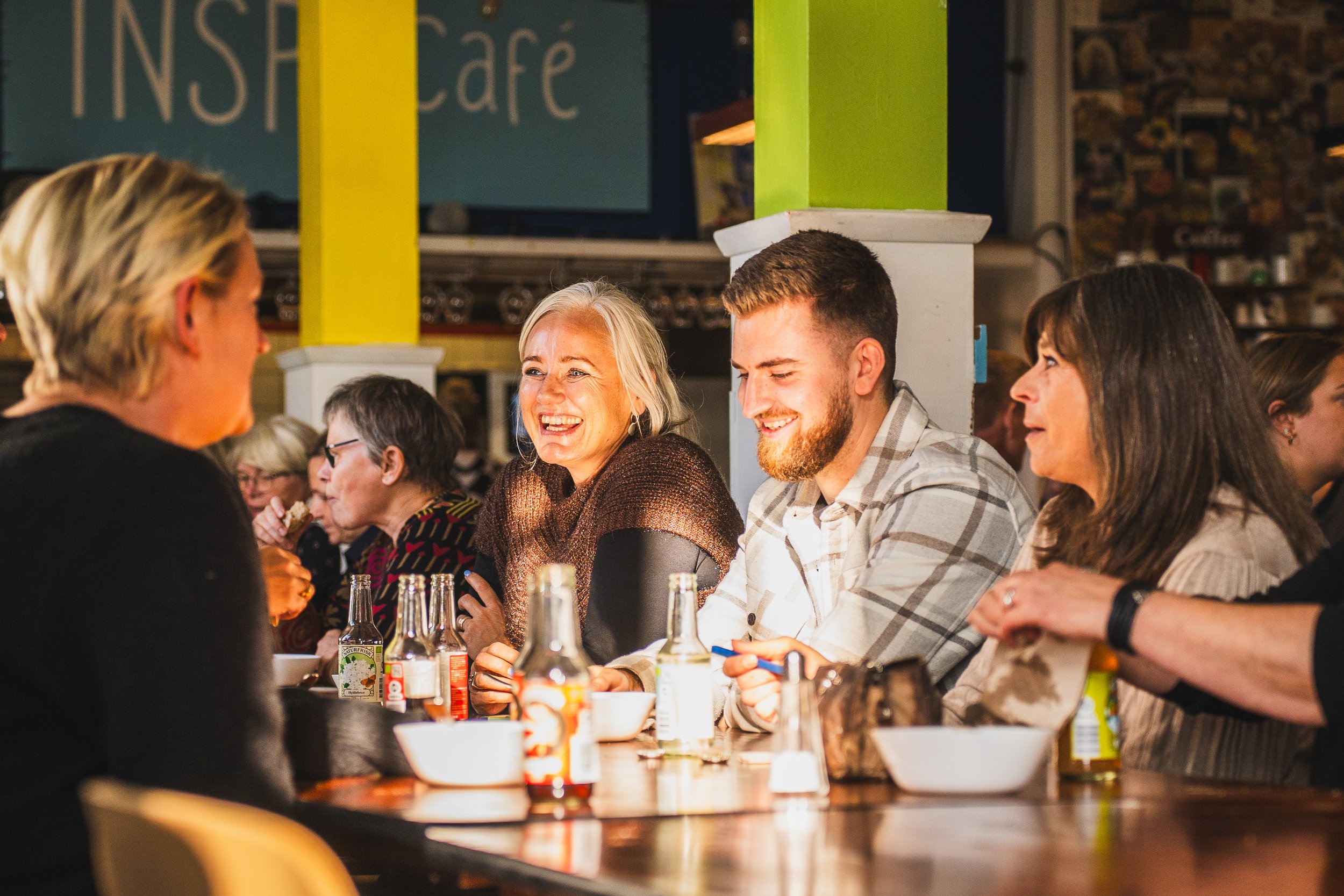 Group of people sitting at a table in a cafe, engaging in conversation, with drinks and food, smiling and enjoying their time.