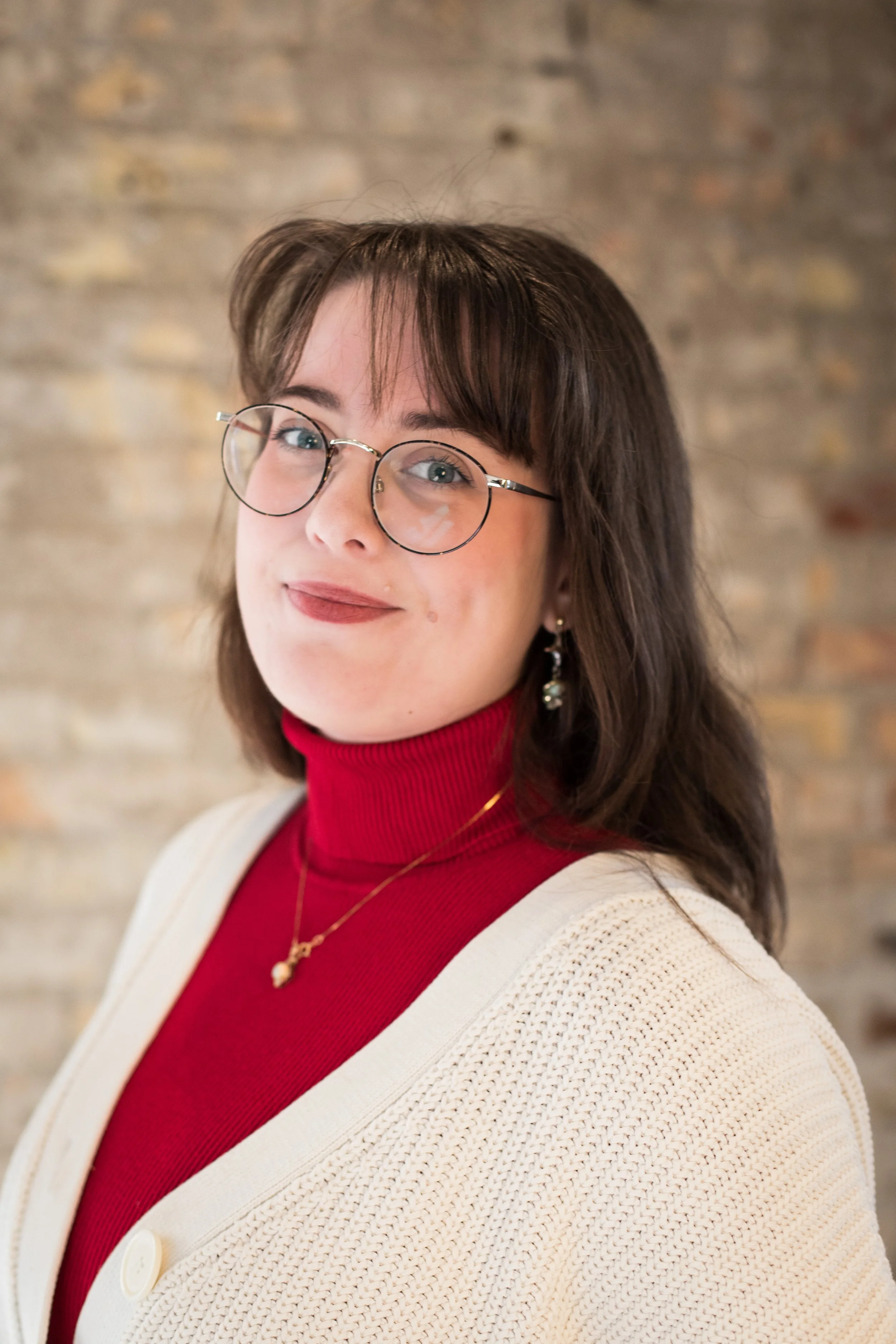 A woman with glasses, wavy brown hair, and a red turtleneck sweater, wearing a cream cardigan and jewelry, smiling at the camera in front of a brick wall.