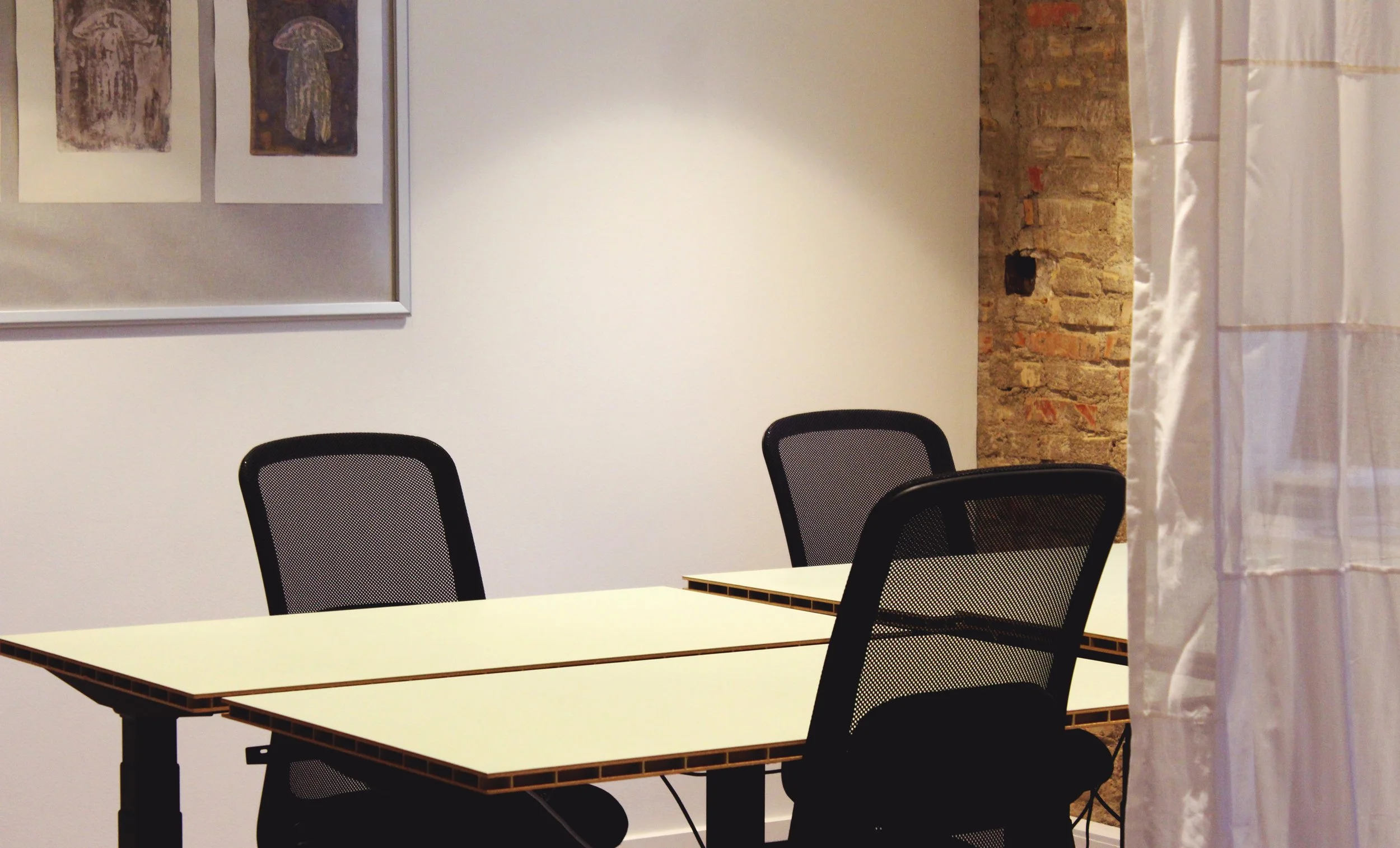 A meeting room with three white tables pushed together, surrounded by three black mesh office chairs. There's a framed artwork on the white wall and exposed brick wall on one side, with a white curtain in the foreground.