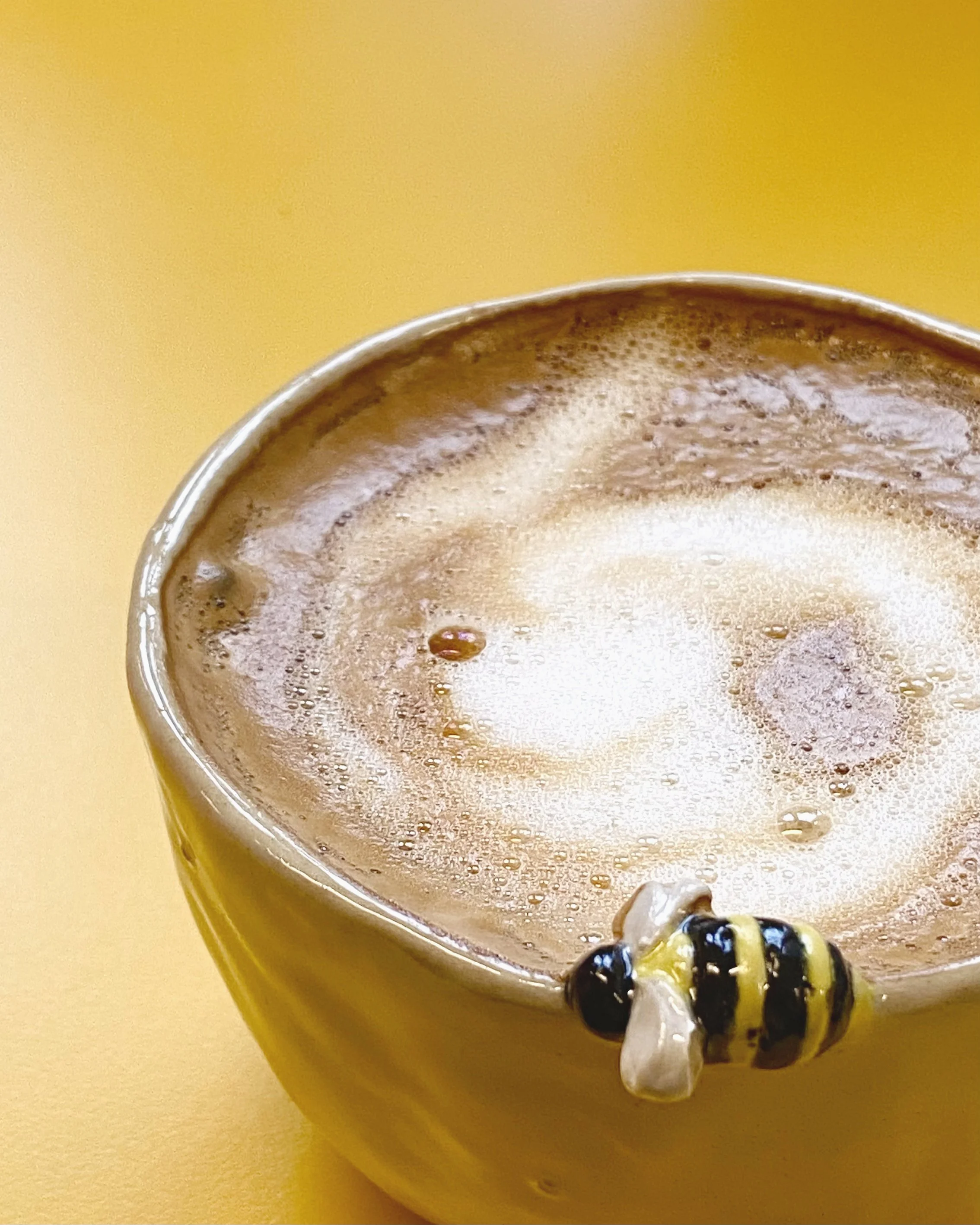 A yellow mug of coffee on top, decorated with a small bee figurine on the rim, against a yellow background.