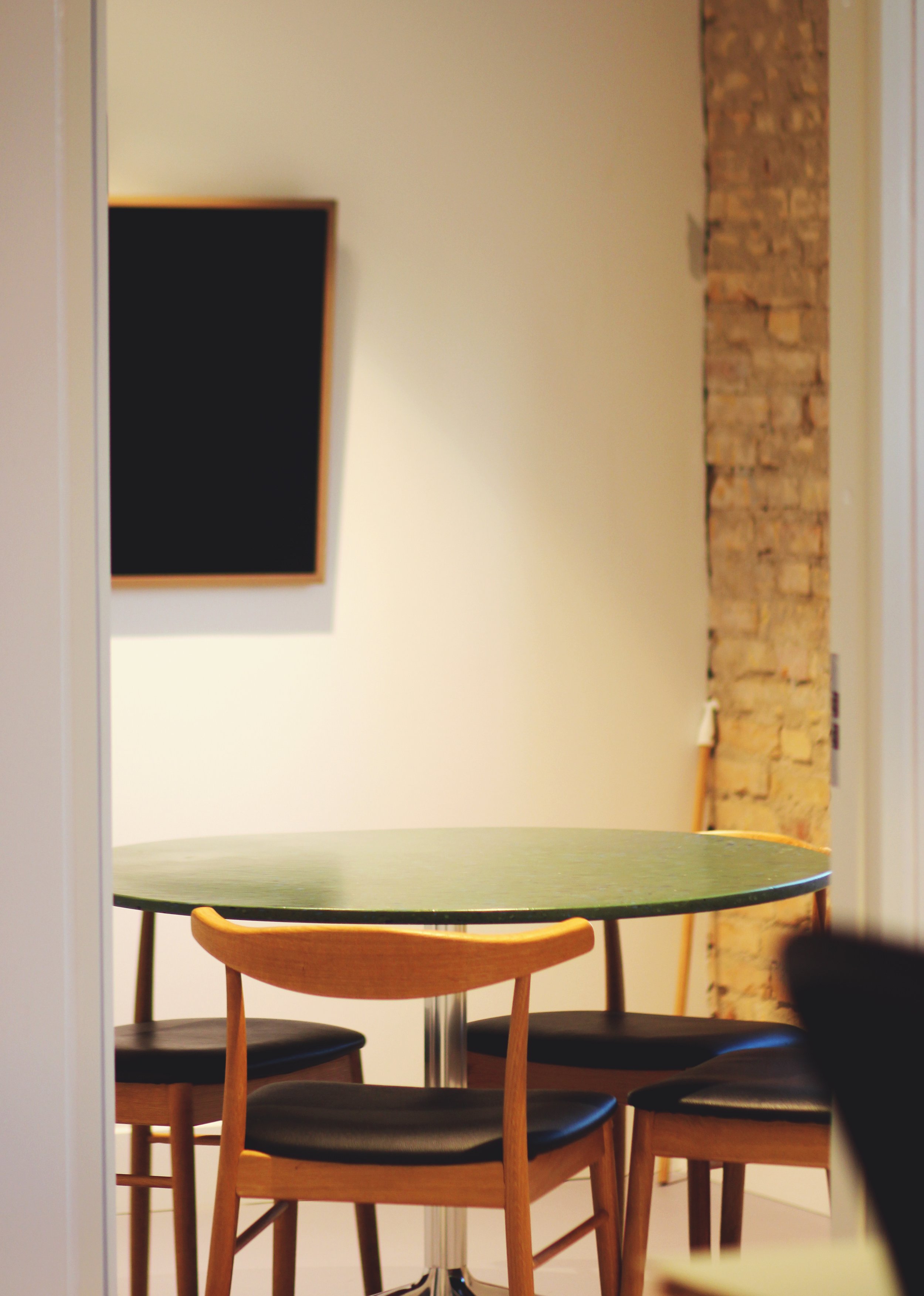 A round green table with four wooden chairs with black cushions, situated in a modern room with a brick wall and a blank blackboard on the wall.