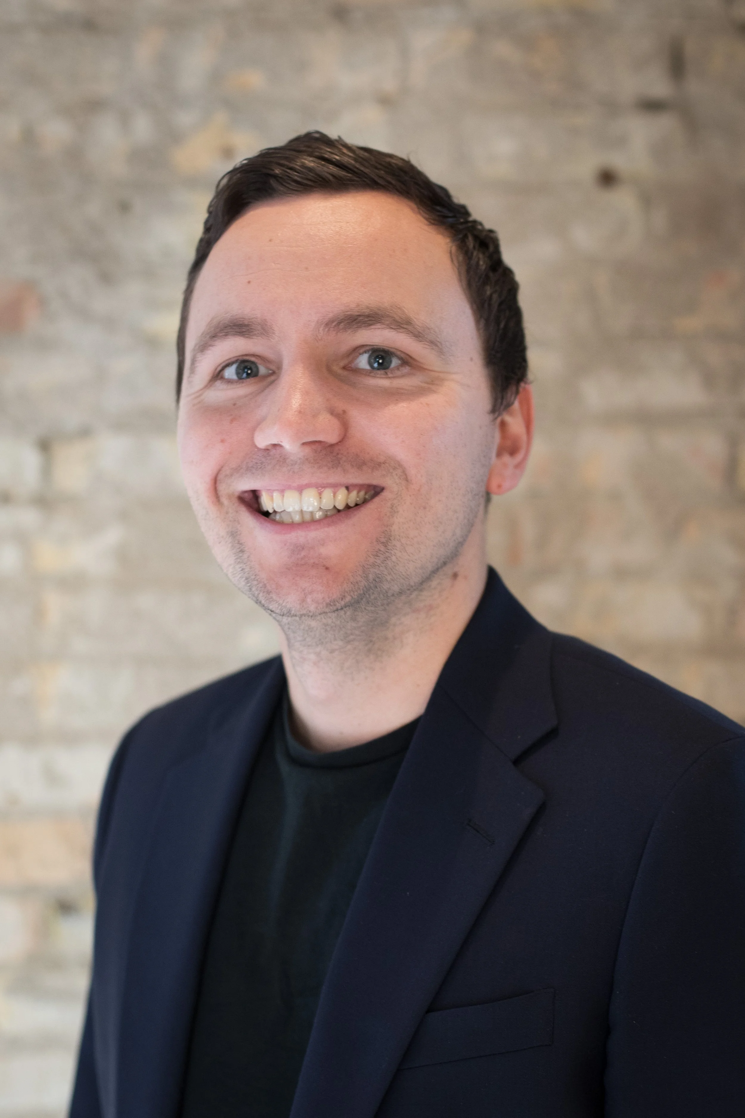 Portrait of a smiling man with short dark hair, light complexion, wearing a black blazer over a black shirt, set against a beige brick wall background.