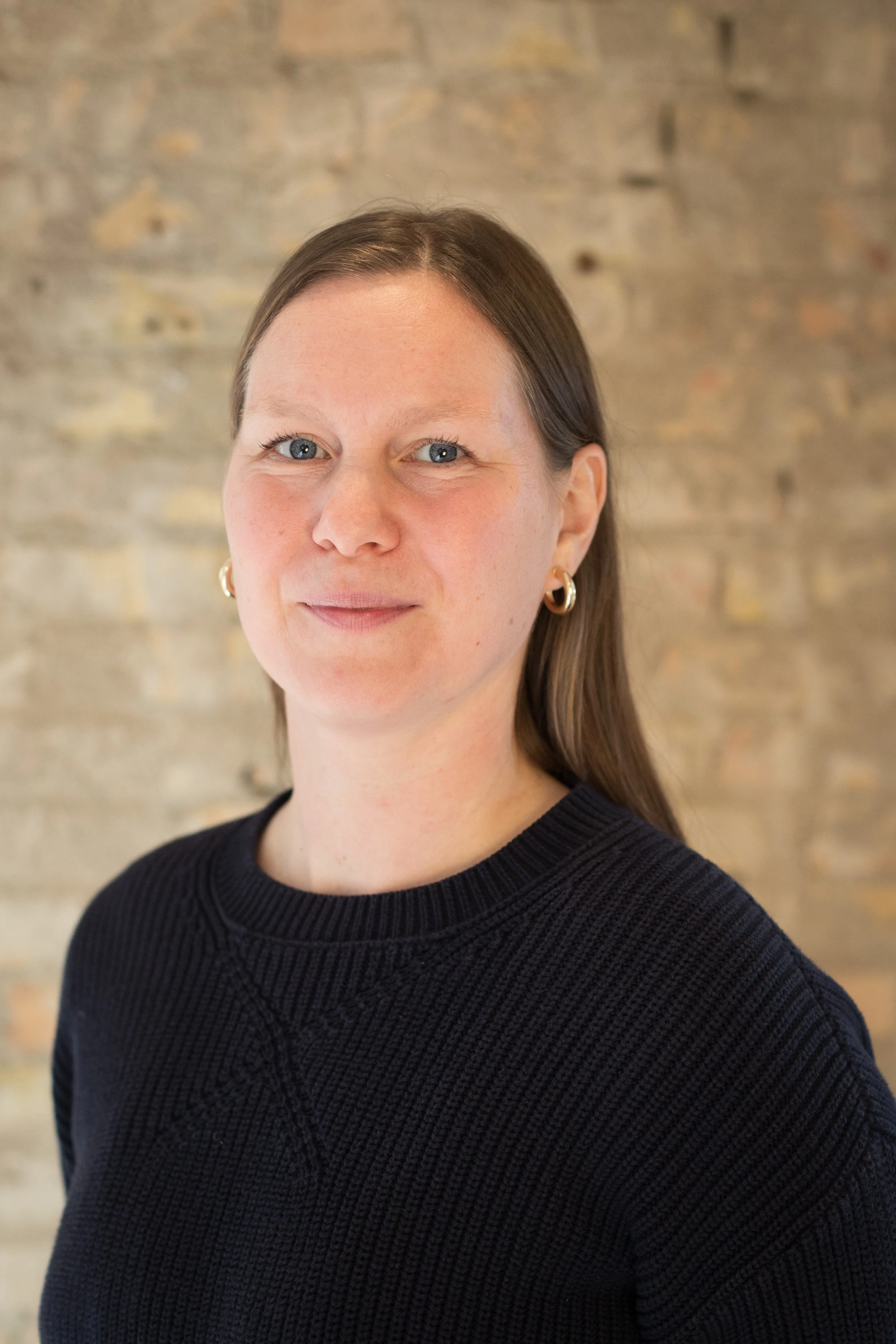 A portrait of a woman with long brown hair wearing a black sweater and gold hoop earrings, standing against a brick wall background.