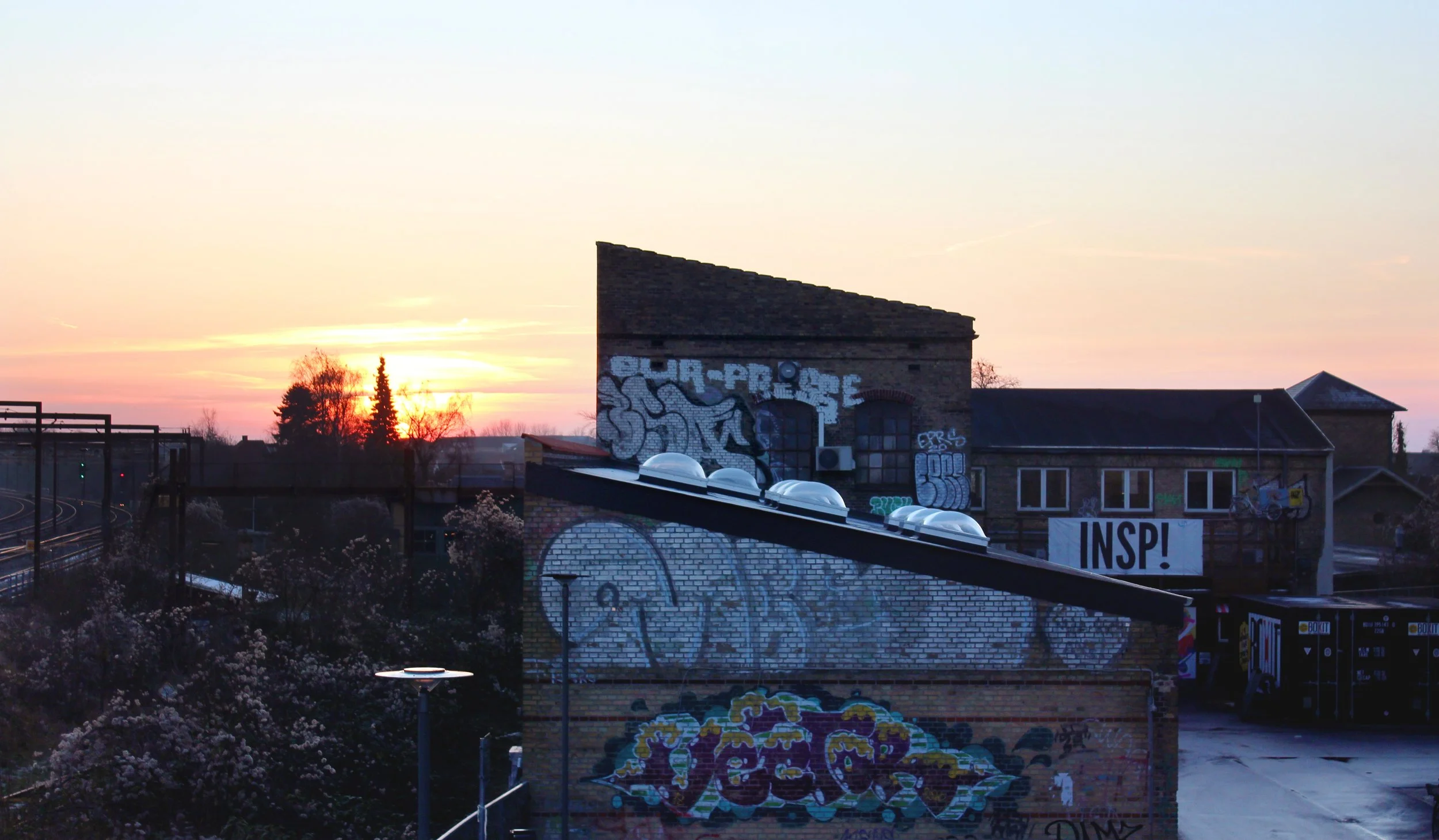Sunset over a graffiti-covered brick building with a sloped roof, next to train tracks and flowering bushes.