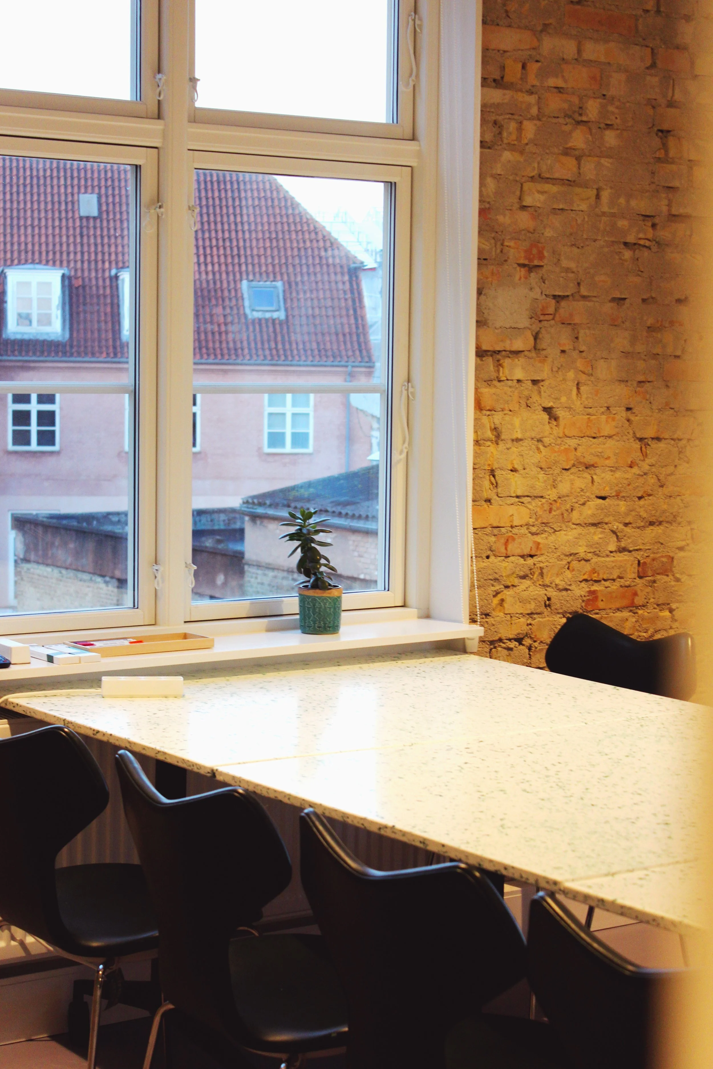 Interior office space with a terrazzo table, black chairs, a window with a small potted plant on the sill, and an exposed brick wall.