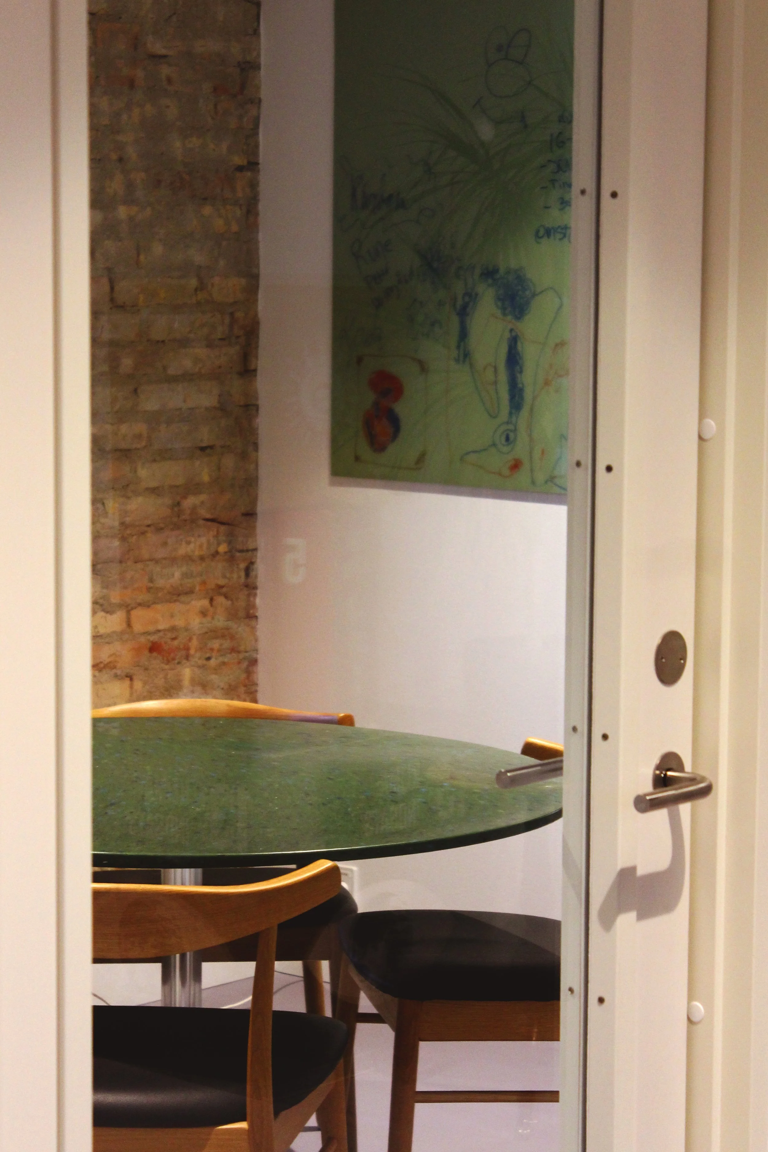 View through a glass door of a room with a round green table and four wooden chairs with black cushions; a whiteboard with colorful drawings and writing is on the wall in the background, and an exposed brick wall is on the left side.