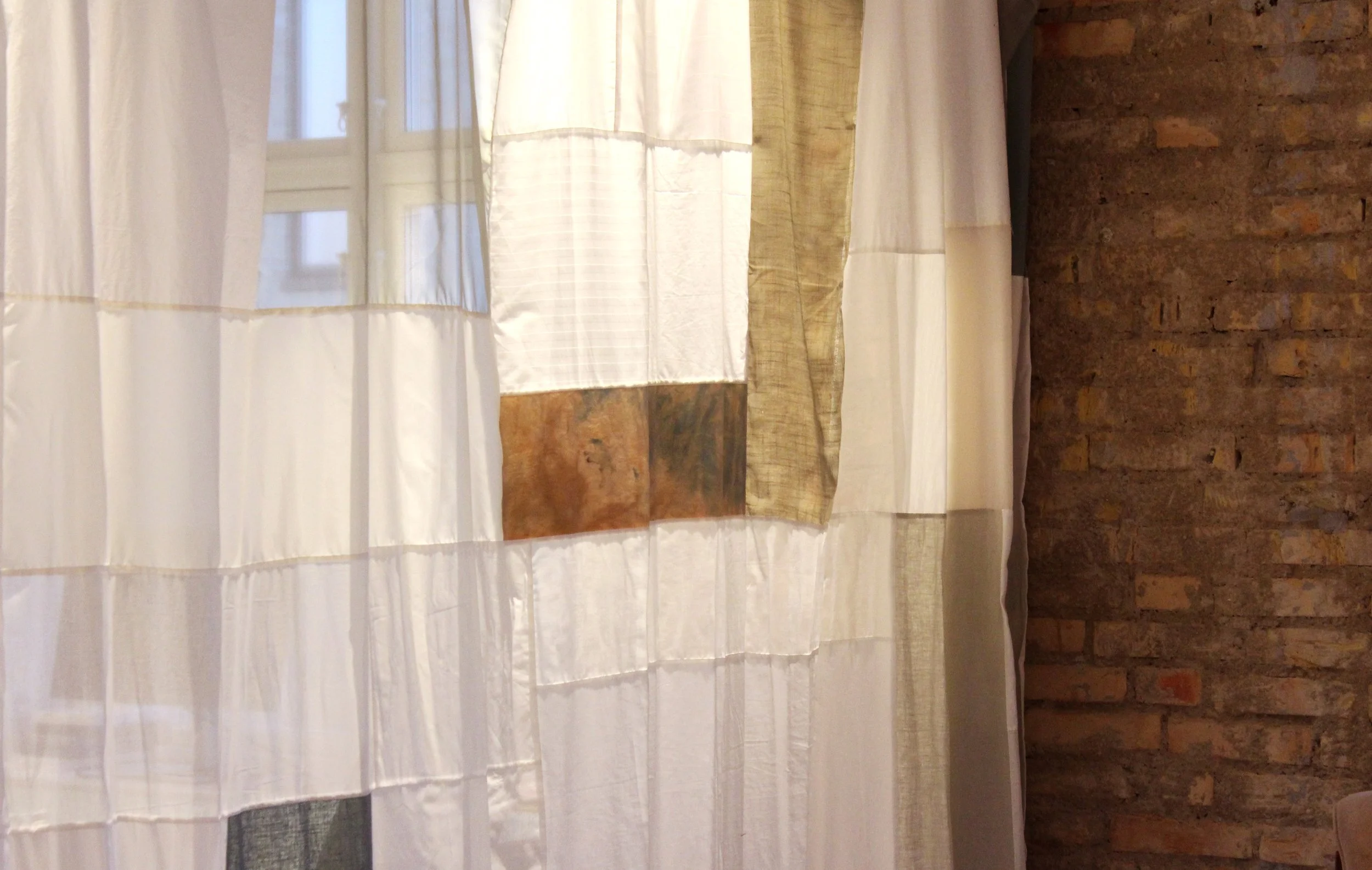 Close-up of a window with white, beige, and light brown patchwork curtains hanging in front of a brick wall.