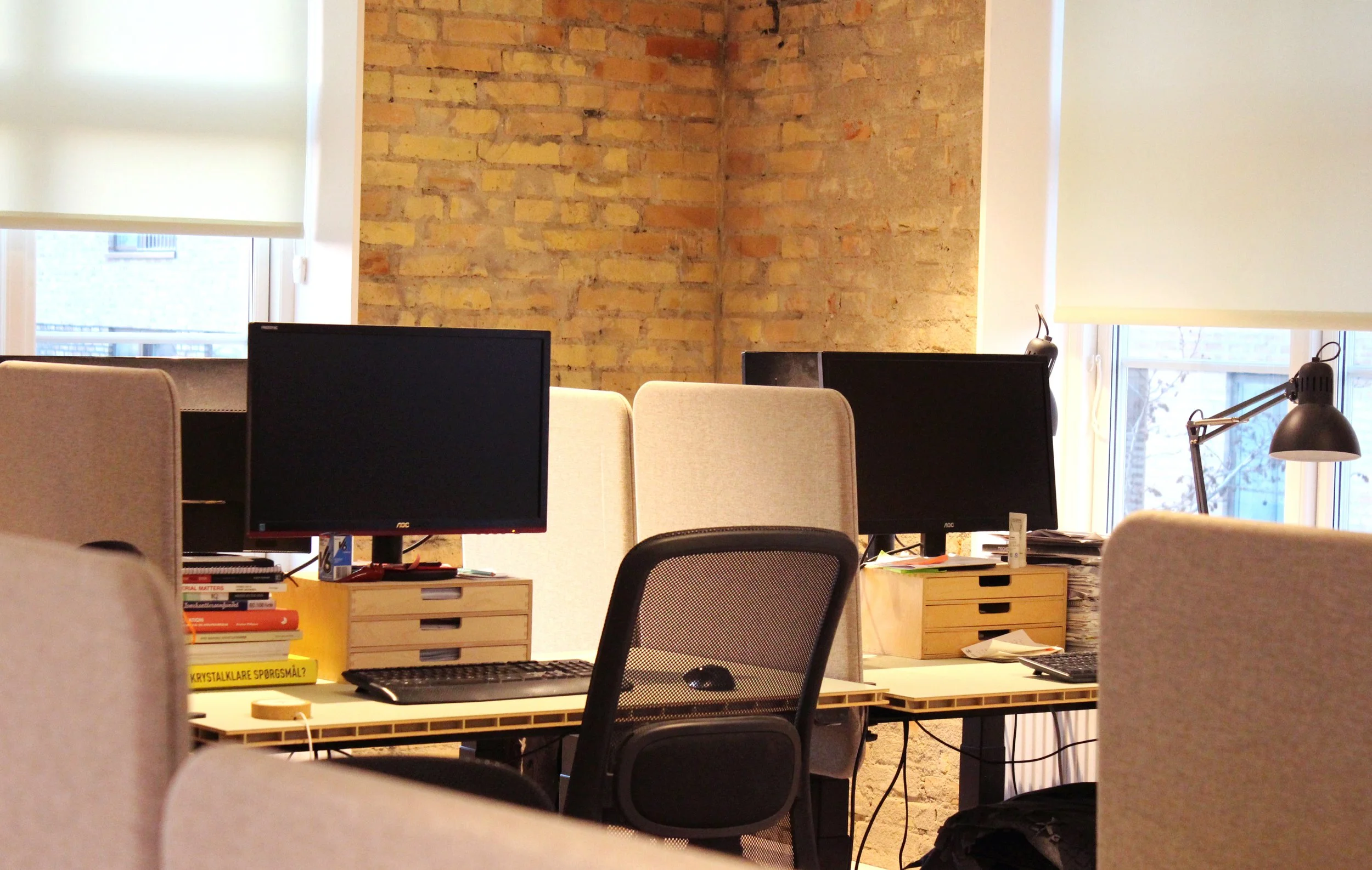 Office workspace with desks, computers, chairs, books, desk organizers, a desk lamp, and large windows with roller blinds, in front of a brick wall