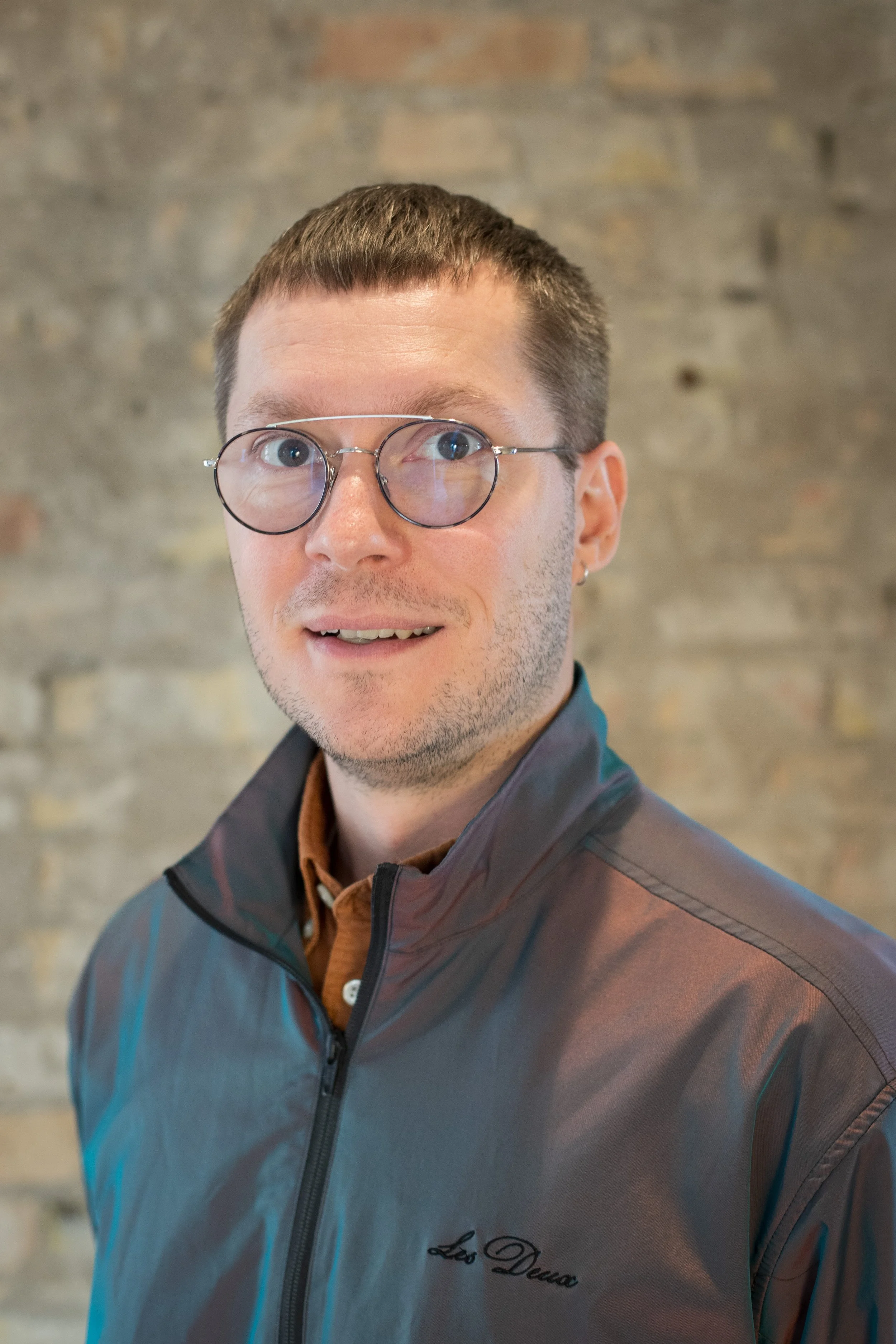 A young man with short brown hair, wearing glasses with round lenses, a light brown shirt, and a gray jacket with a logo that says 'Leo Dena,' stands in front of a brick wall.