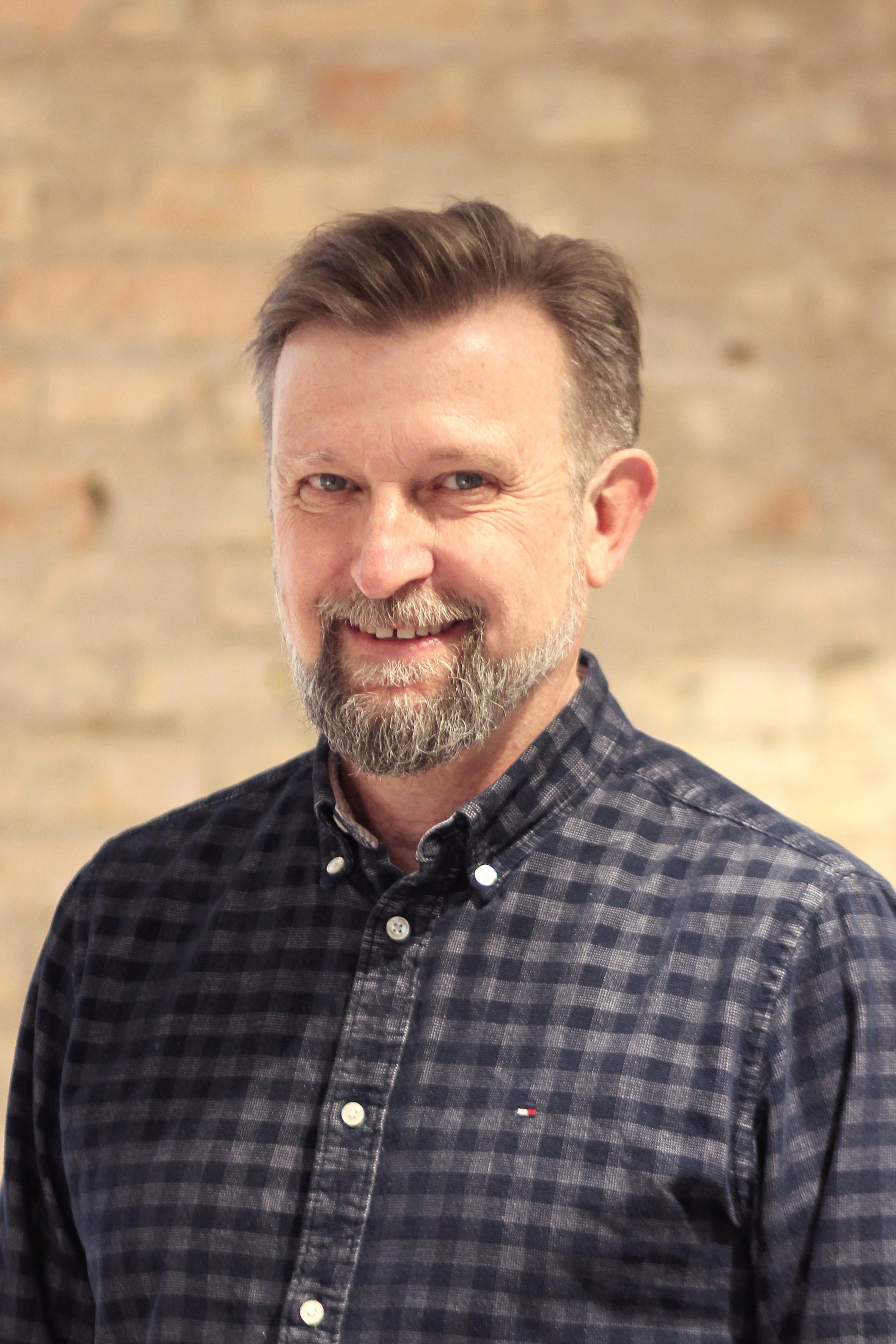A smiling middle-aged man with a beard and short hair wearing a black and gray checkered button-up shirt, standing against a light beige brick wall background.