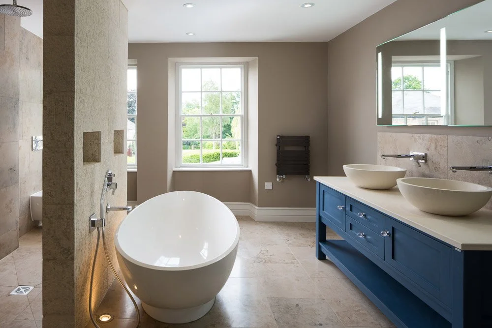 one point perspective of bathroom with large dark blue double sink unit, freestanding curved bath and glimpse into shower, pale beige tiles on floor and walls