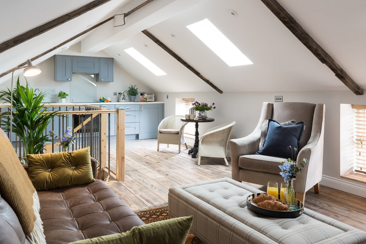  Loft room holiday let living space with sky lights,, brown leather sofa in foreground, arm chair and breakfast table along far wall, on back wall powder blue kitchen 