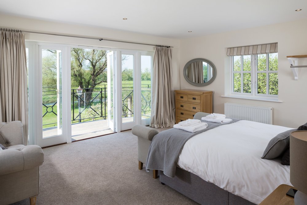  bright wide angle image of bedroom with beige carpet, French doors leading to balcony overlooking river and countryside  