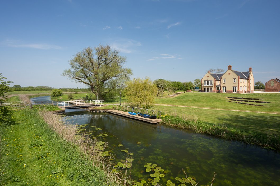  elevated wide angle image showing river, lawn and house with a small bridge leading over the river, house is surrounded by countryside 