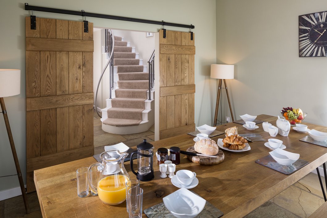 bright image of breakfast table set up with pastries, juice and coffee, looking through sliding doors towards curved staircase  