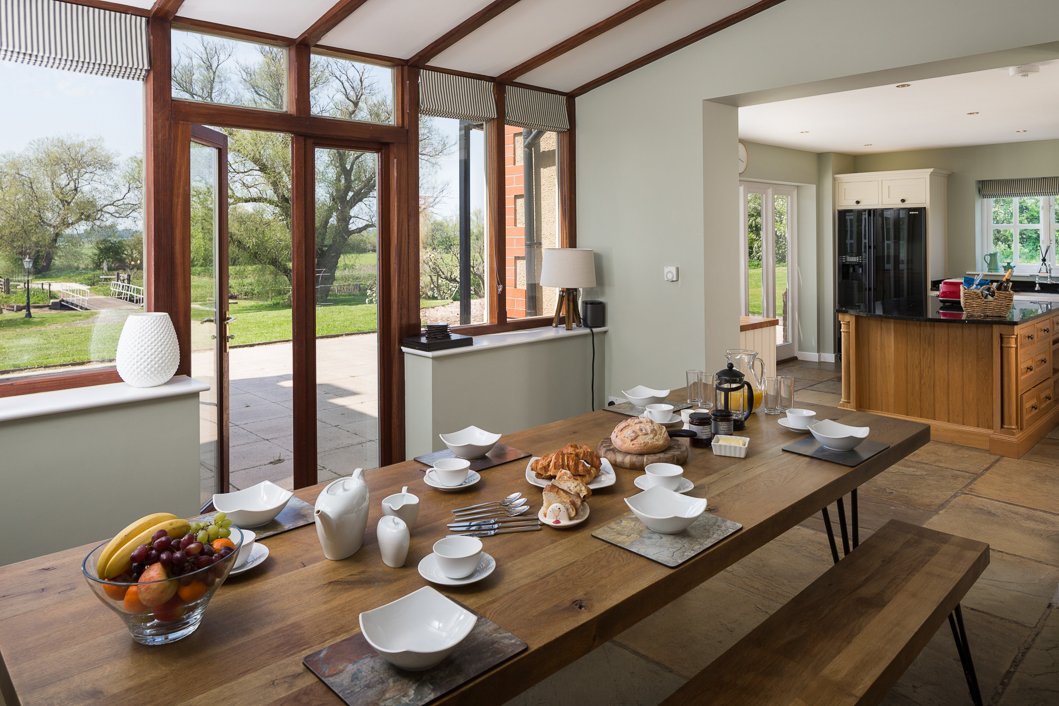  bright image looking over breakfast table set up with pastries, juice and coffee looking out towards garden and into open plan kitchen  