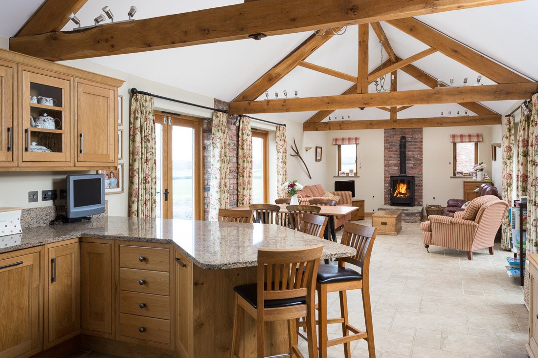  well lit, long kitchen/ family room with tall lofted beamed ceilings, wooden units, striped sofas and log burner 