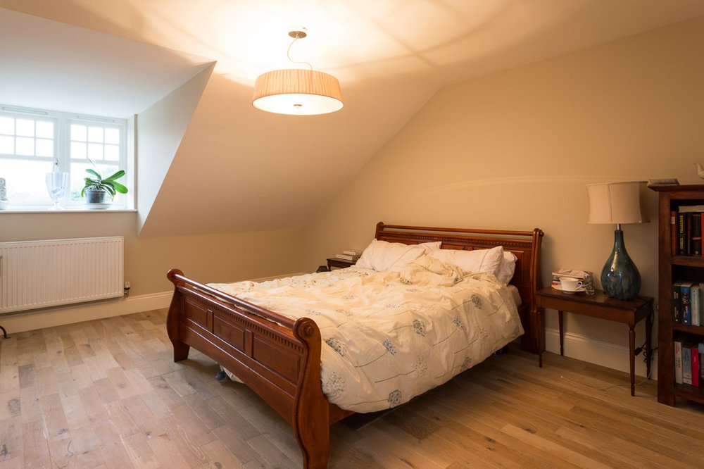  same bedroom with wooden sleigh bed, pale wooden flooring, white walls badly lit and messy with bedsheets not tidy, very orange ceiling light 