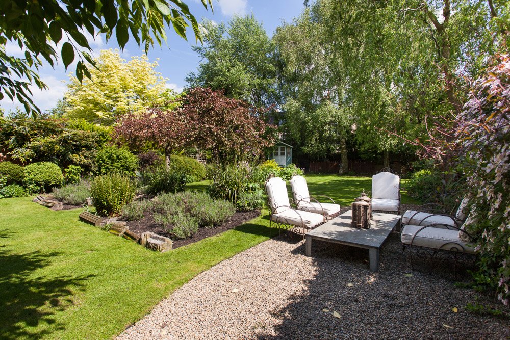  sunny garden with mature trees and shrubbery, small gravel area with garden furniture   