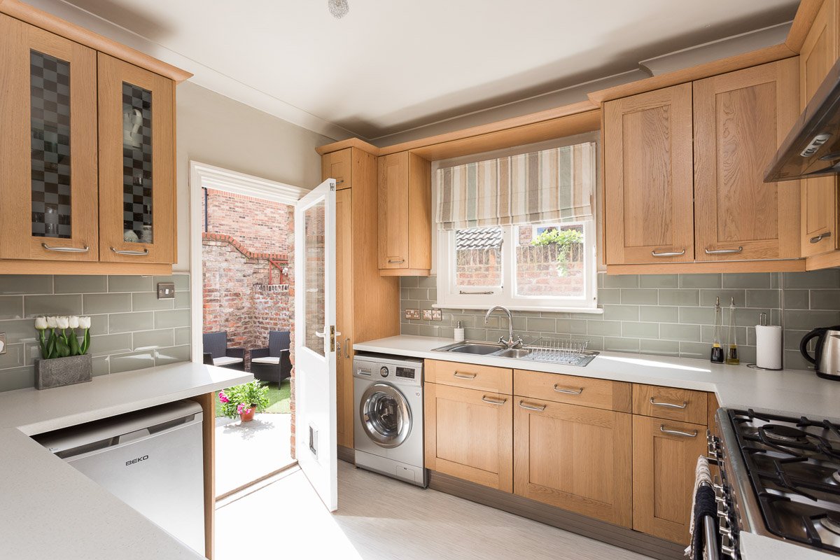  image of kitchen with wooden units, pale flooring, open door leading to garden 