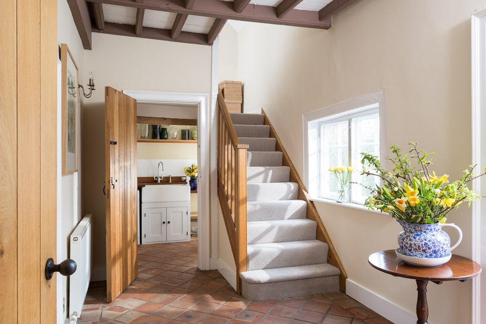  well lit farmhouse hallway with red tile flooring, cream walls, glimpse into under stairs utility, grey carpet stairs 